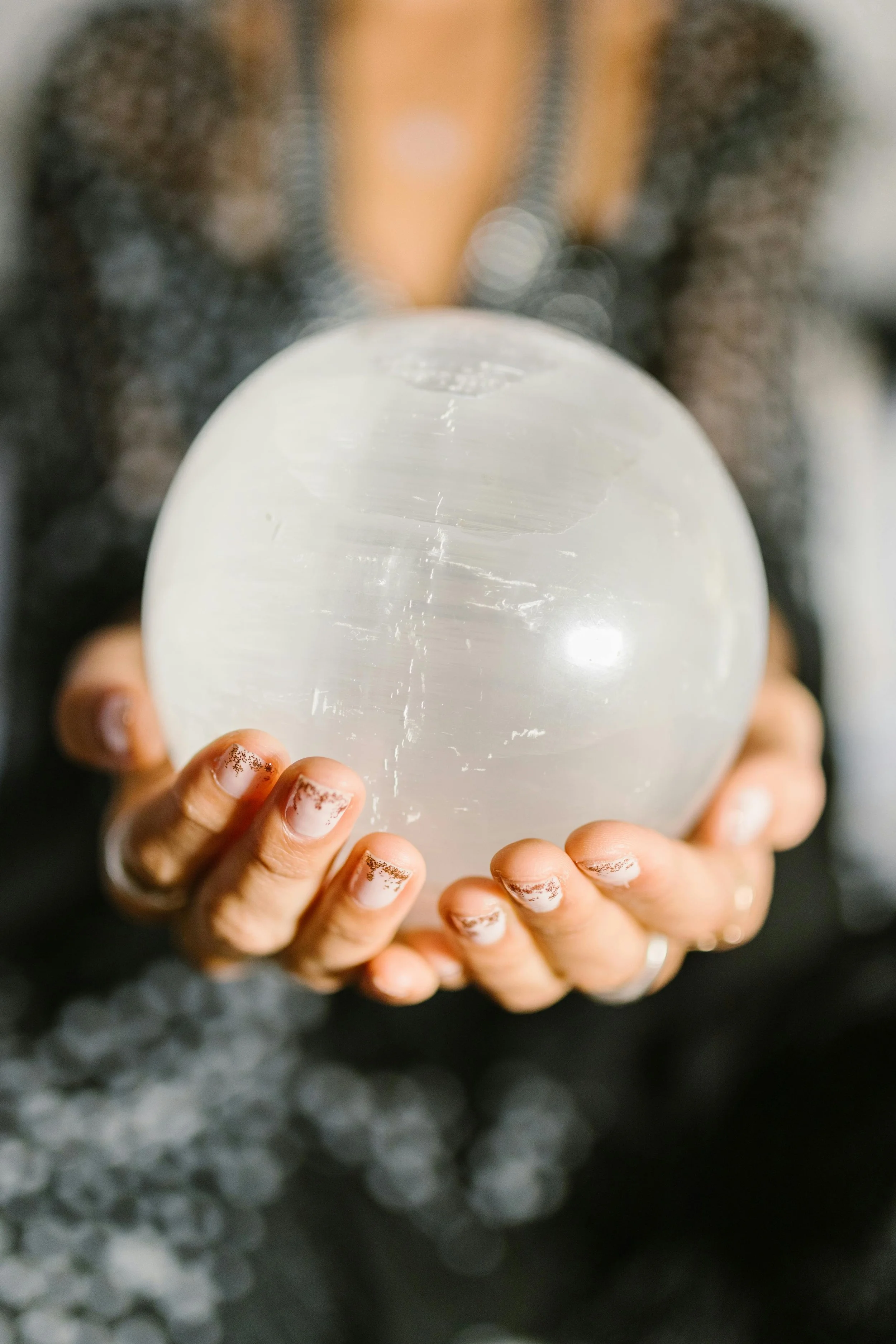 Person holding a glowing selenite crystal ball with painted nails and blurred background.