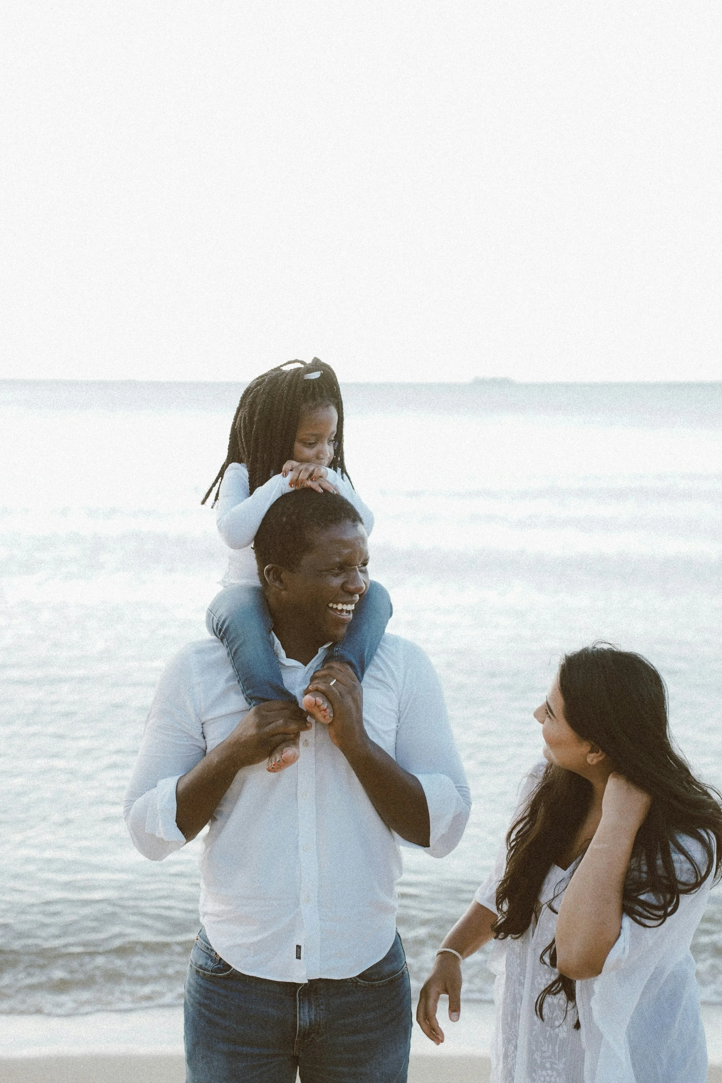 A family enjoying a day at the beach, with a man carrying a girl on his shoulders, and a woman standing beside them, all smiling.