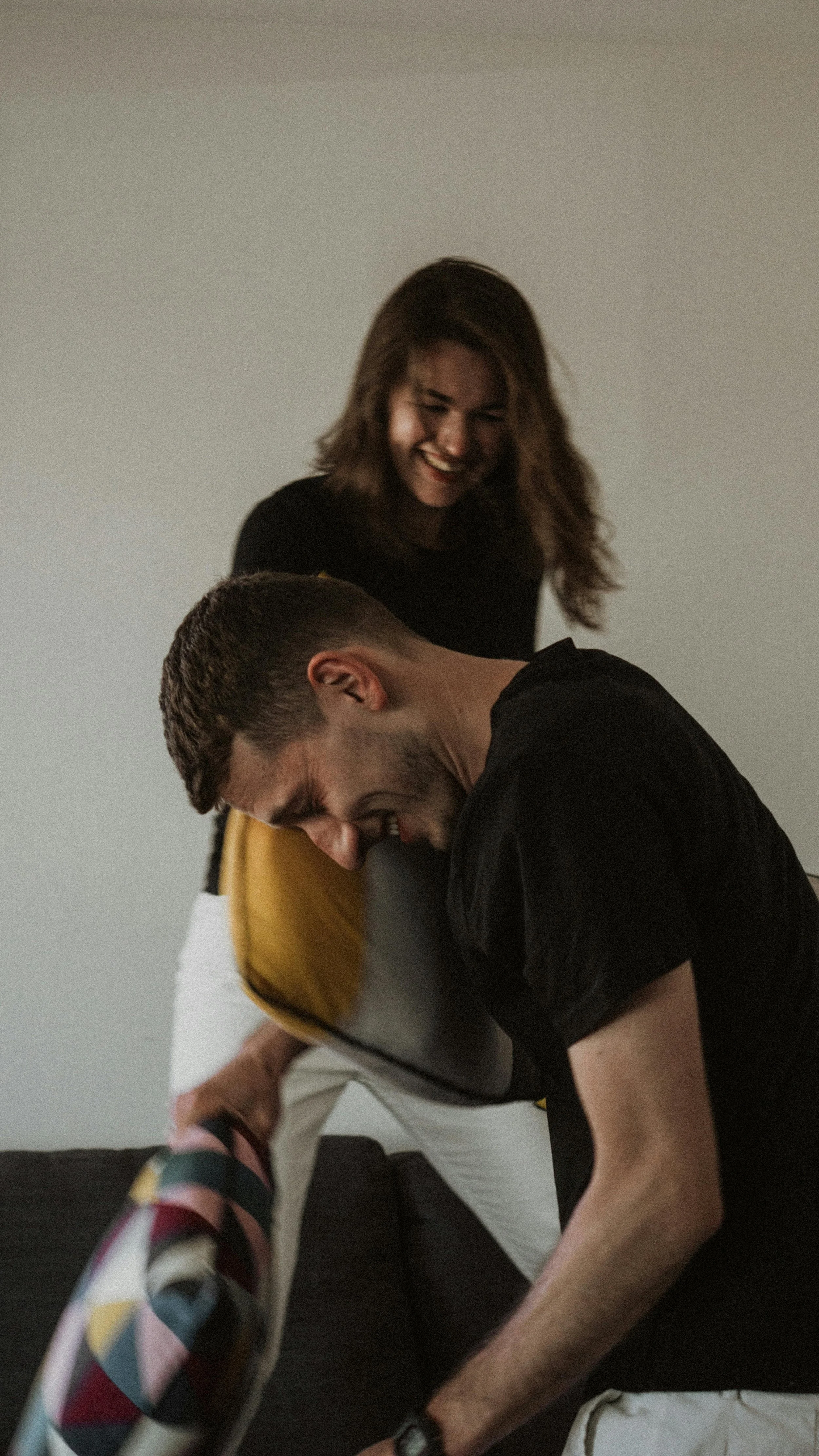 A man and a woman are laughing and having fun, with the man leaning over a couch while the woman stands behind him, all enjoying a lighthearted moment.