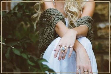 Close-up of a woman with long blonde hair, wearing a gray sweater, resting her hands on her knees, with rings on her fingers, outdoors surrounded by greenery.