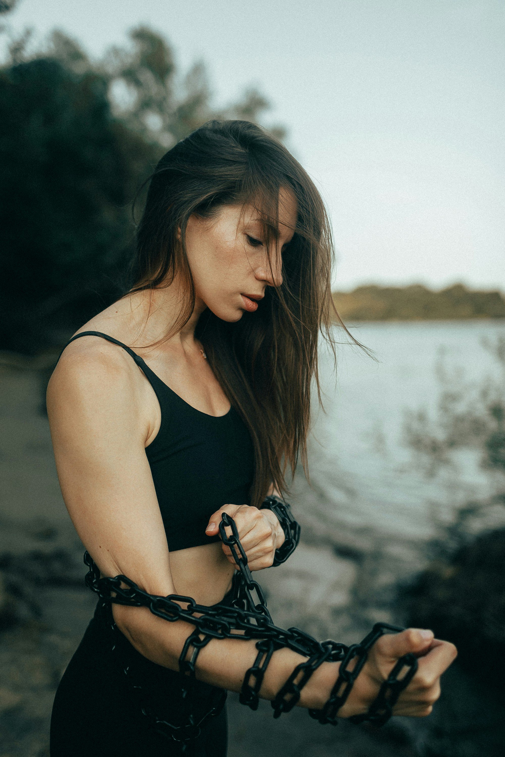 A woman with long brown hair, wearing a black sports bra, stands outdoors near water. She is wrapped in a black chain that coils around her arms and torso, with her head bowed and a contemplative expression.