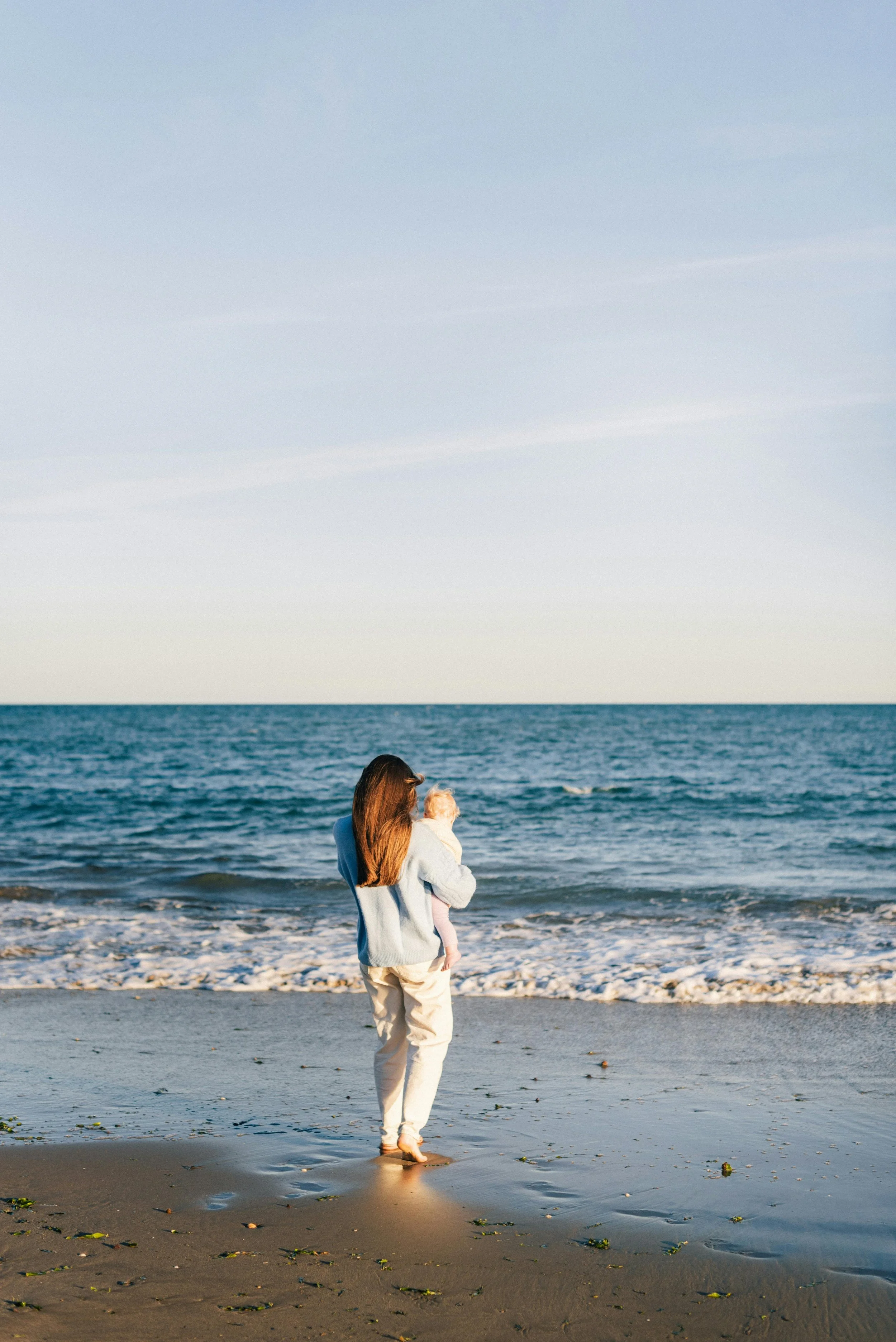 A woman standing on a sandy beach holding a small dog, looking at the ocean during sunset.