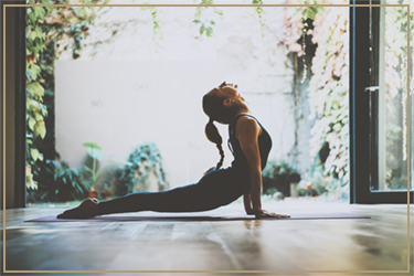 A person practicing yoga in a lunge pose on a wooden floor, with sunlight and greenery visible through an open door