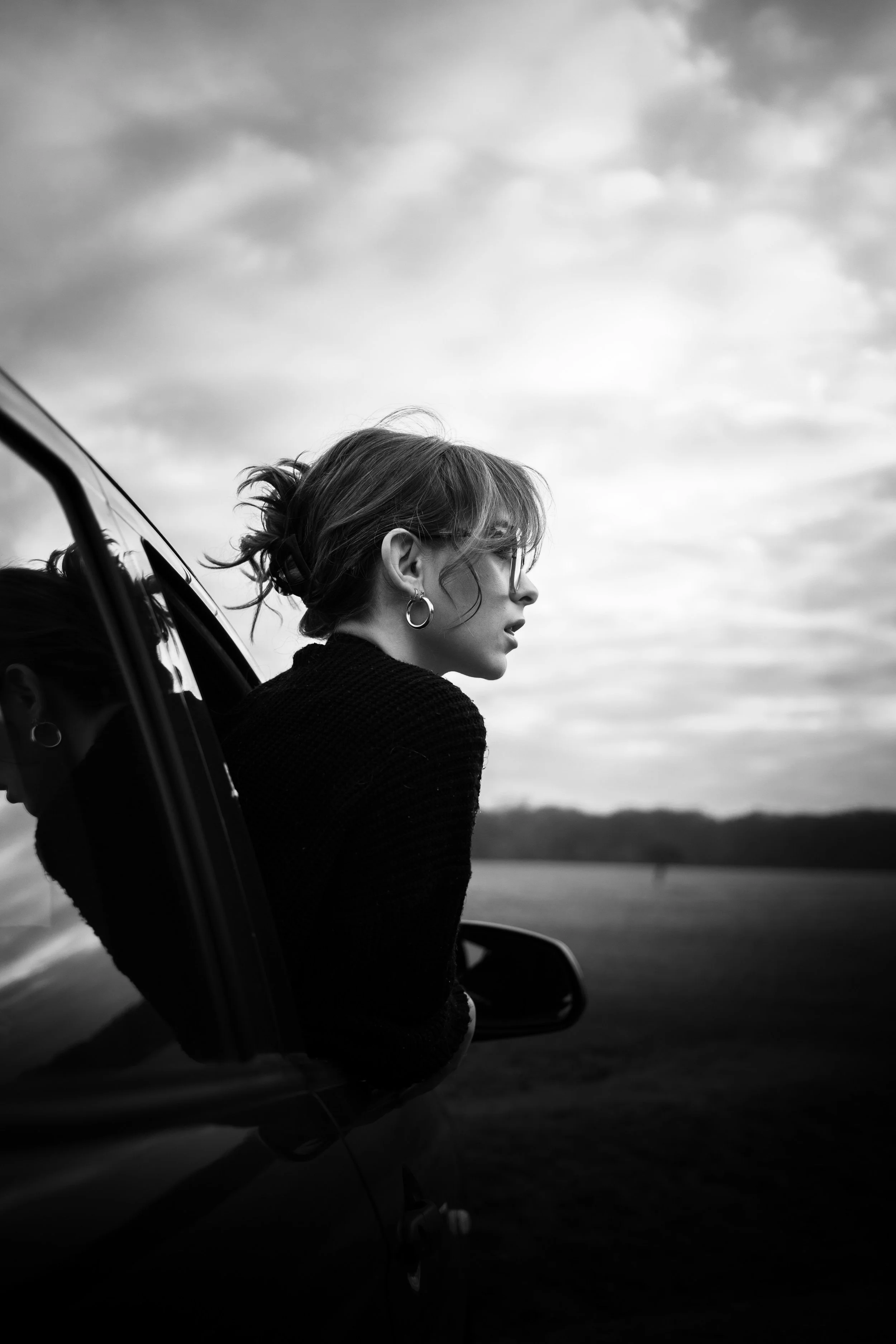 A woman leaning out of a car window, looking to the side, with a cloudy sky and open field in the background. The photo is in black and white.