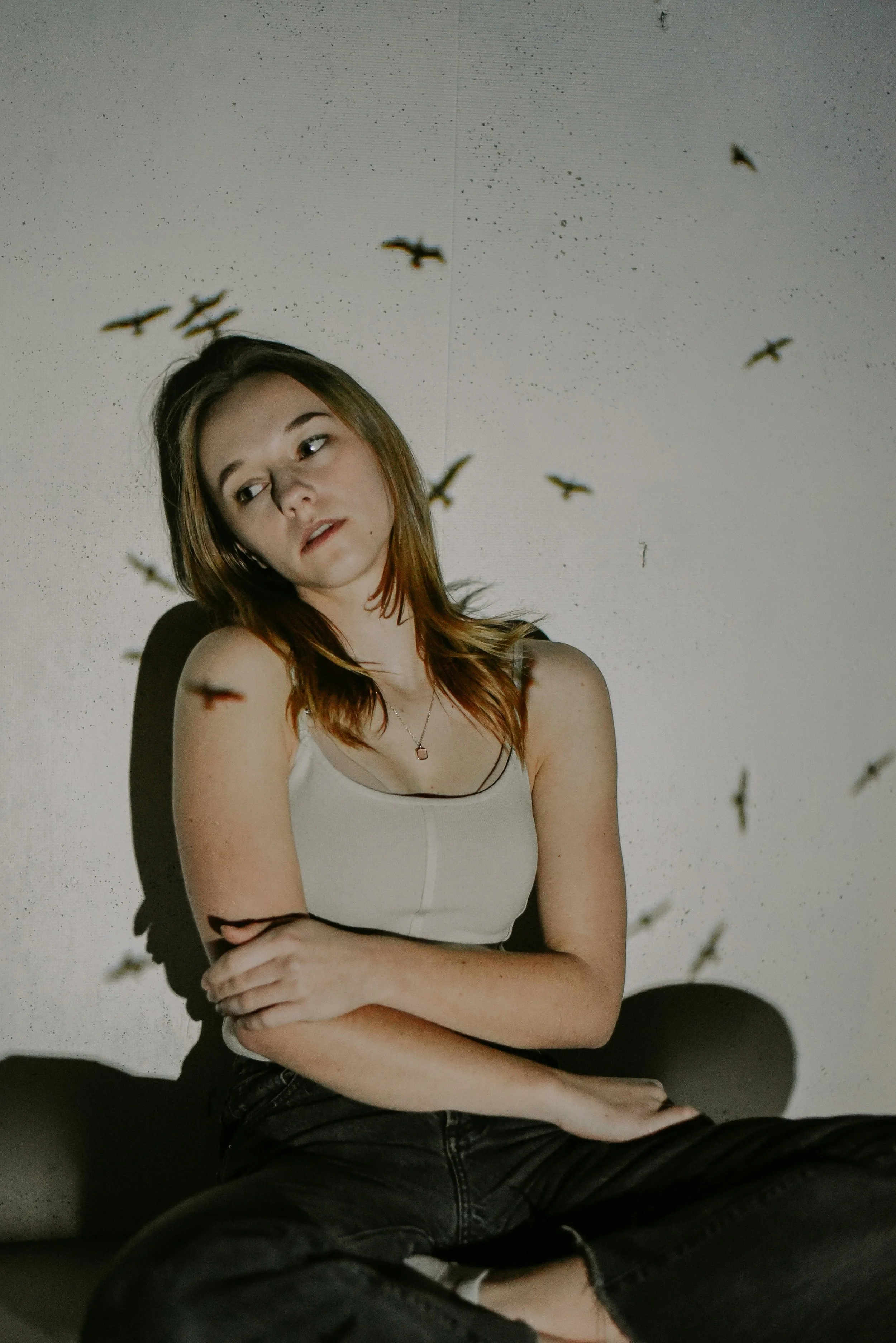 Young woman sitting against a plain wall with bird shadows projected onto the wall around her.