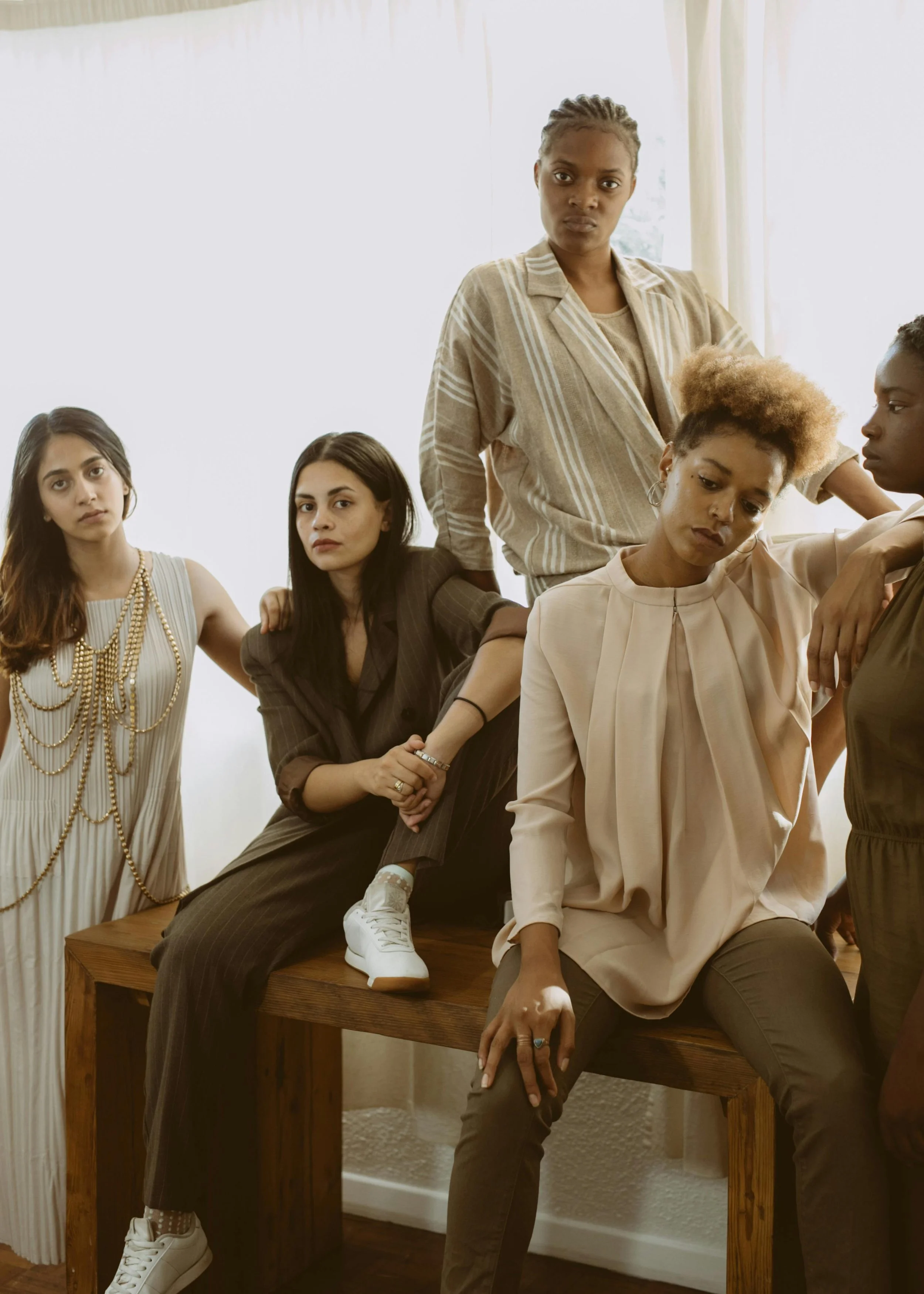 A group of five women with diverse skin tones posing together indoors, dressed in stylish clothing with serious expressions.
