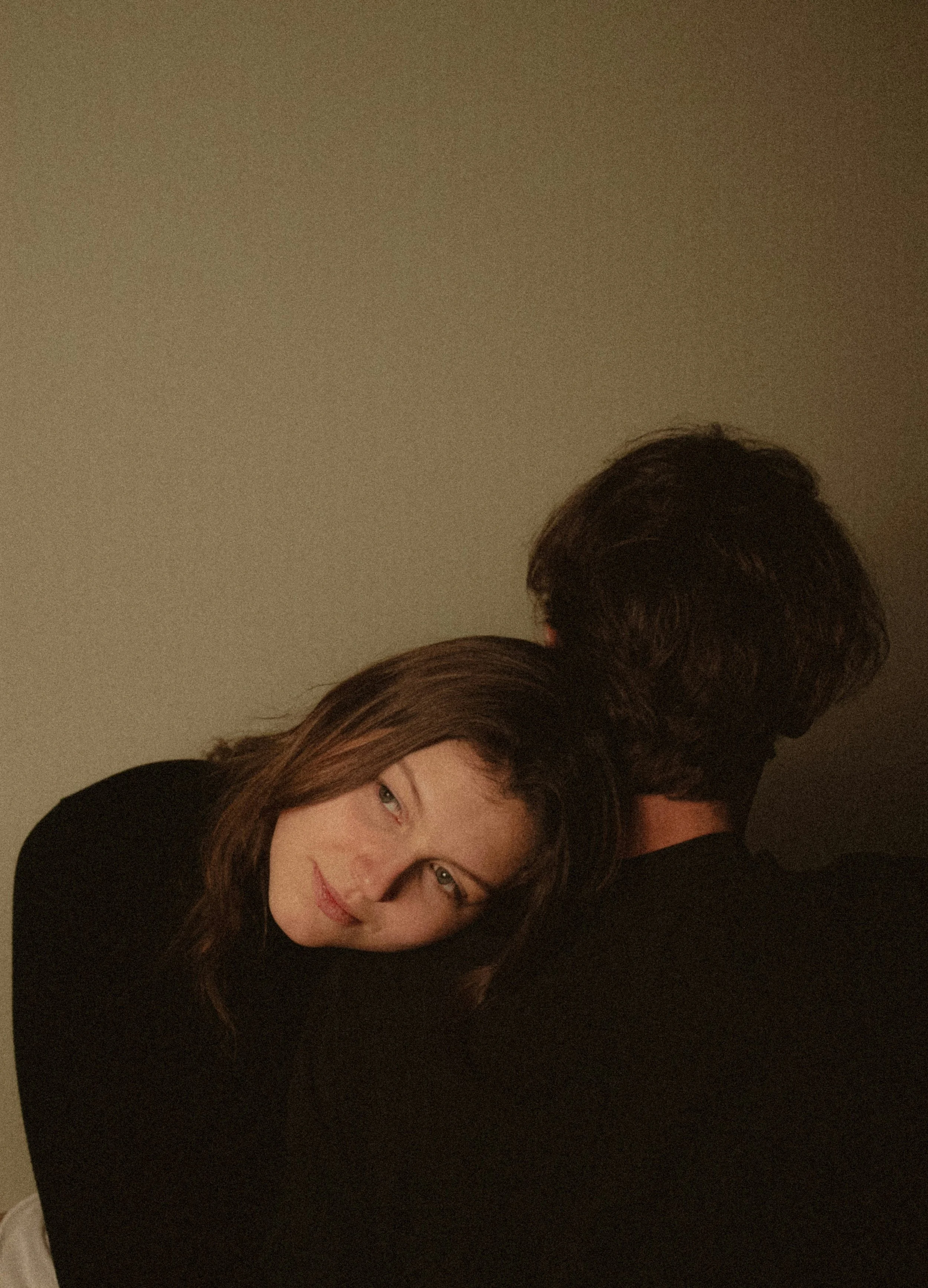 A woman with light brown hair resting her head on a man's shoulder, facing the camera with a gentle smile, against a neutral background.