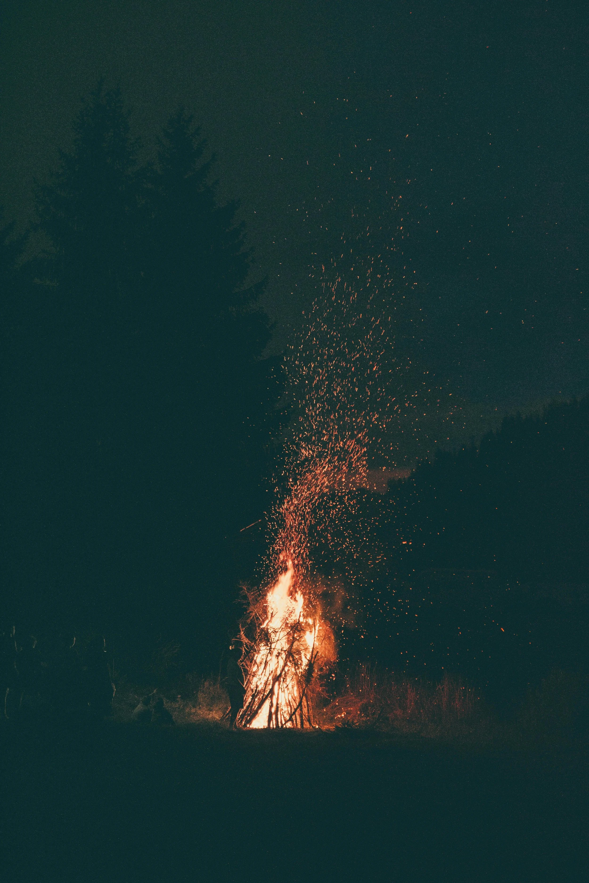 A large campfire with flames and sparks rising into the night sky, surrounded by trees.
