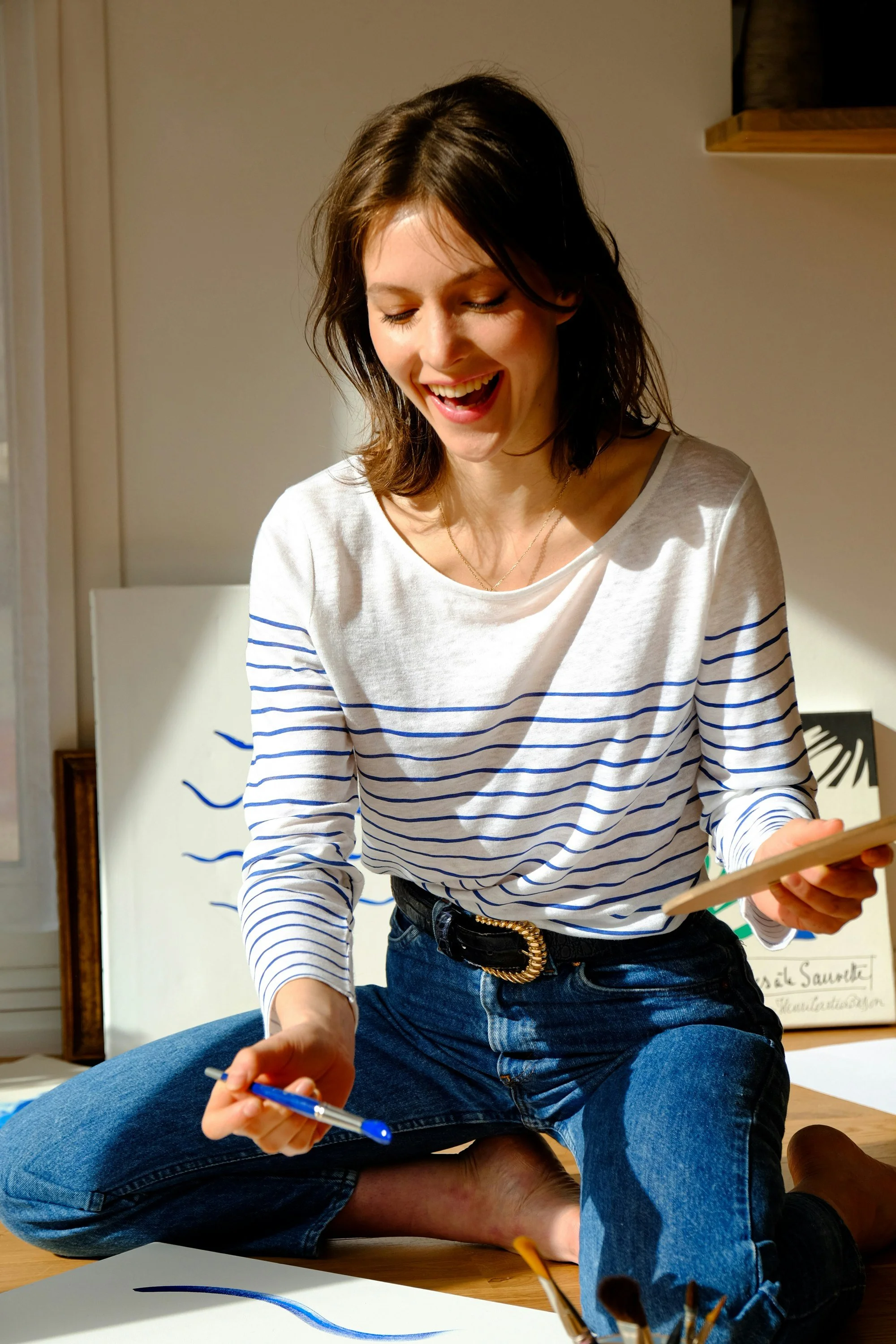 A young woman with shoulder-length brown hair, sitting on the floor, smiling while holding a blue pen in one hand and a tablet in the other, with artwork and art supplies around her.