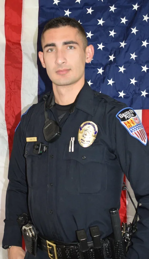 A woman with dark, curly hair smiling, wearing glasses, hoop earrings, and layered necklaces, in a blurred indoor background.