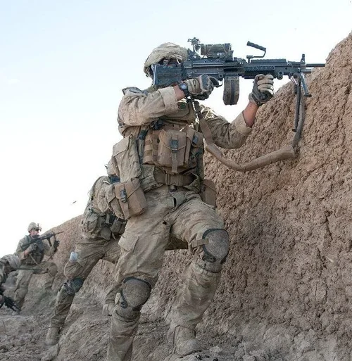A man with curly dark hair, glasses, and a beard, wearing a dark green sweater and a white shirt underneath, looking slightly to the side with a thoughtful expression. In the background, a blurred plant and wall are visible.