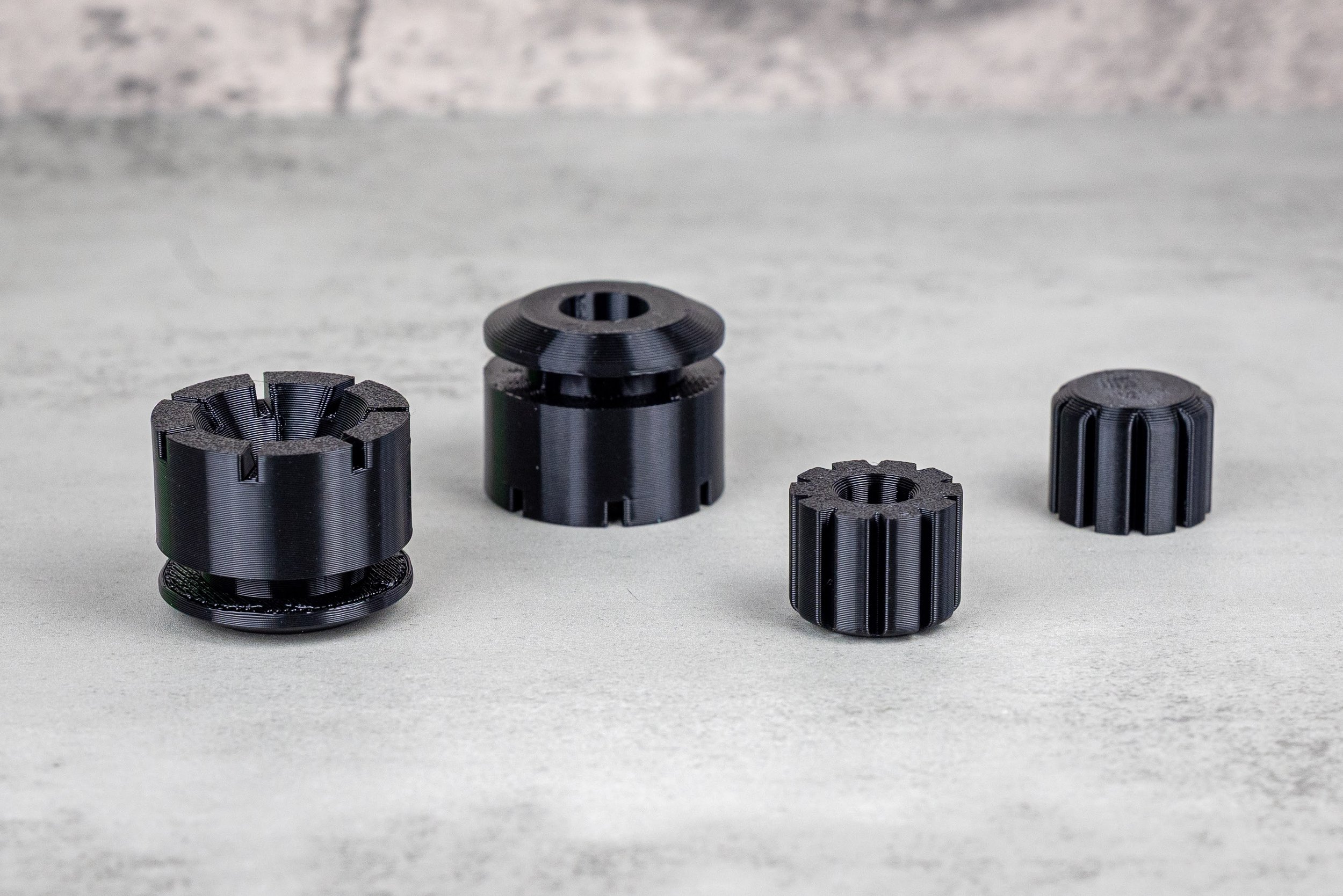 Four black 3D-printed cylindrical mechanical parts with slotted sides and threaded openings on a gray surface