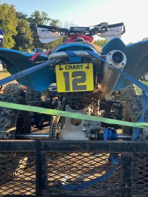 Rear view of a blue off-road ATV with a yellow number 12 plate secured on a utility trailer.