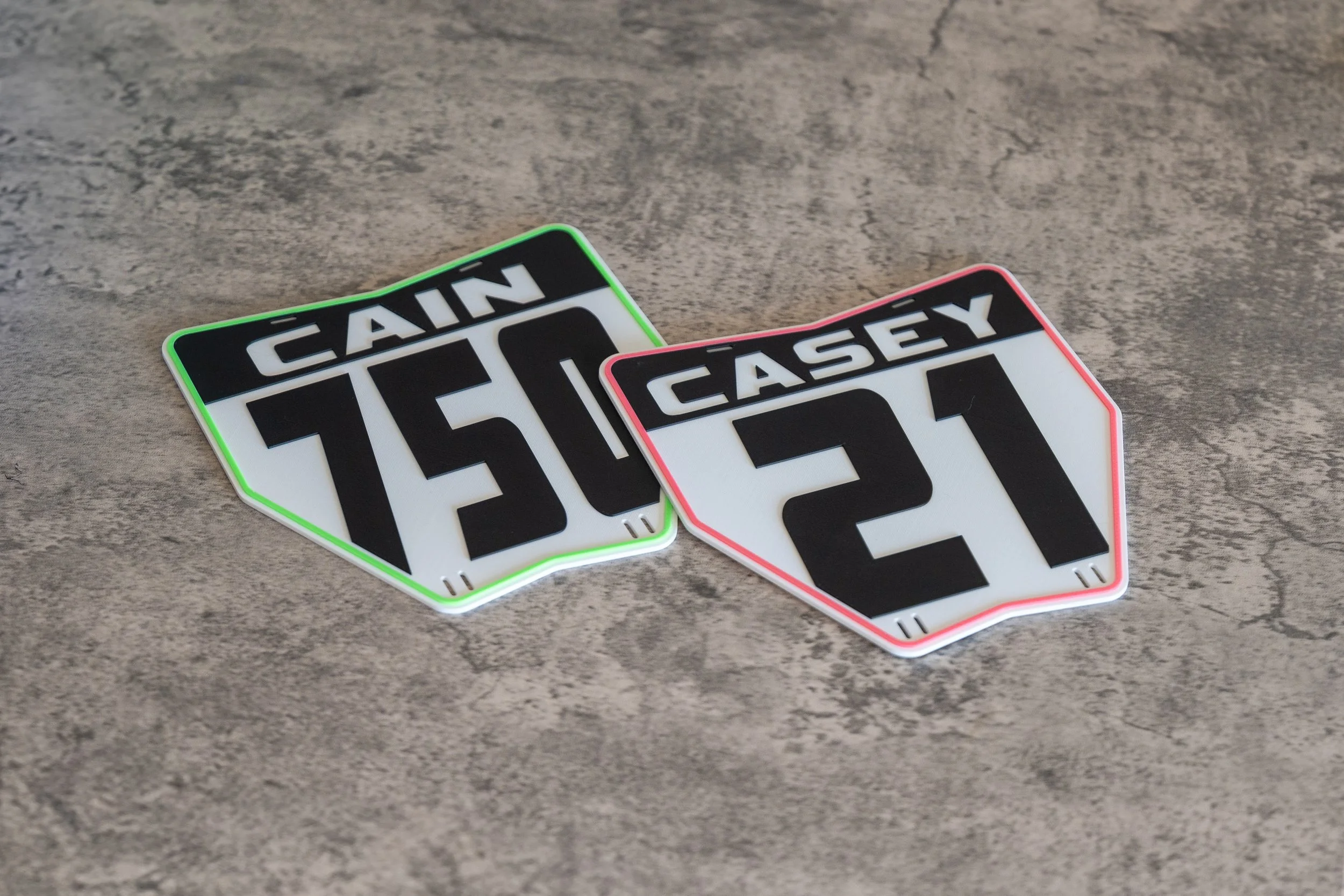 Two overlapping custom stacyc motorcycle number plates labeled “RAIN” and “EASEY” on a gray wooden surface.