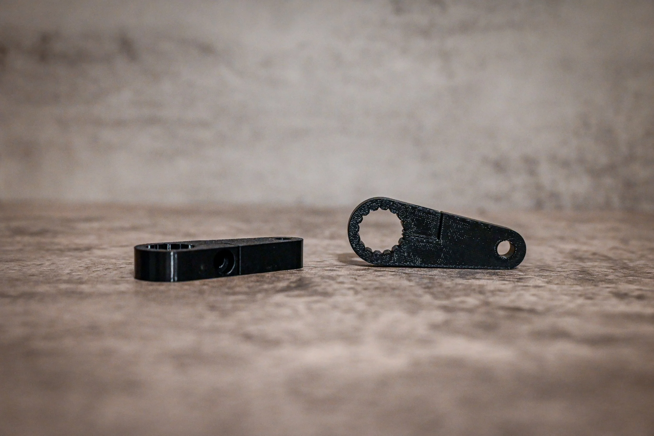 Two black number plate brackets on a wooden surface, including a toothed key-shaped tool and a small bracket.