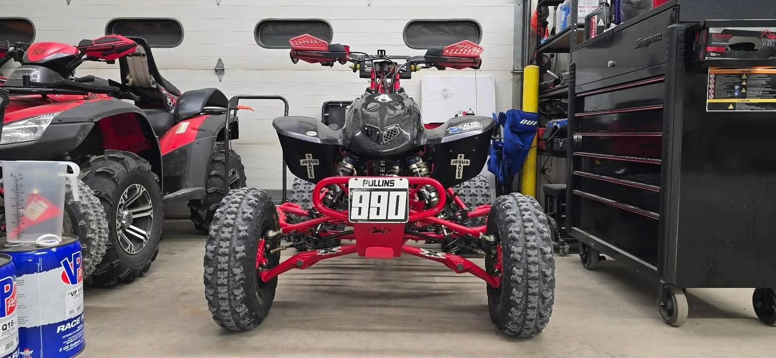 Front view of a customized red and black racing ATV in a garage, with a second ATV, tool chest, and racing supplies.