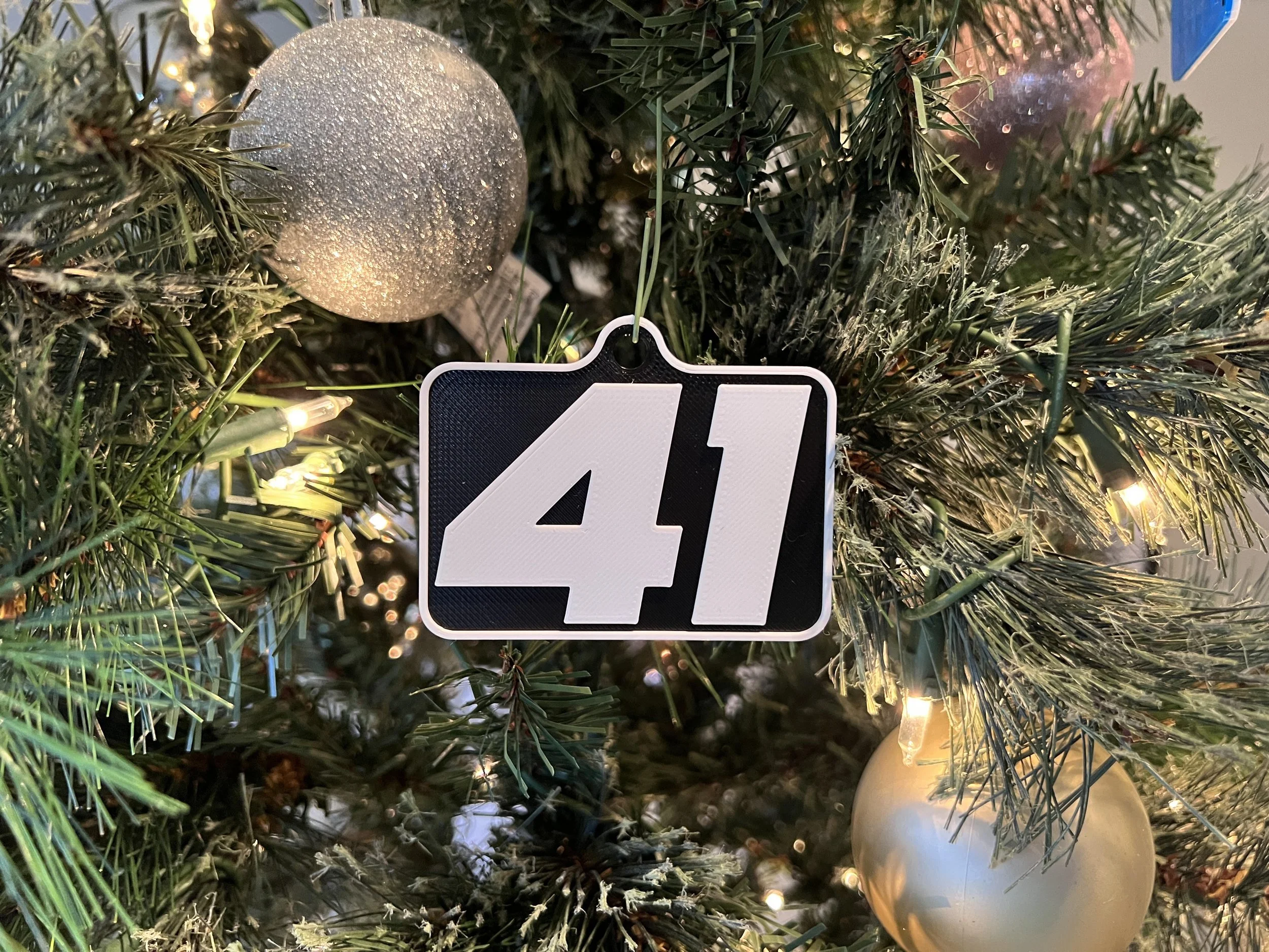 Close-up of a decorated Christmas tree with warm string lights, silver ornaments, and a black-and-white custom ornament number plate.