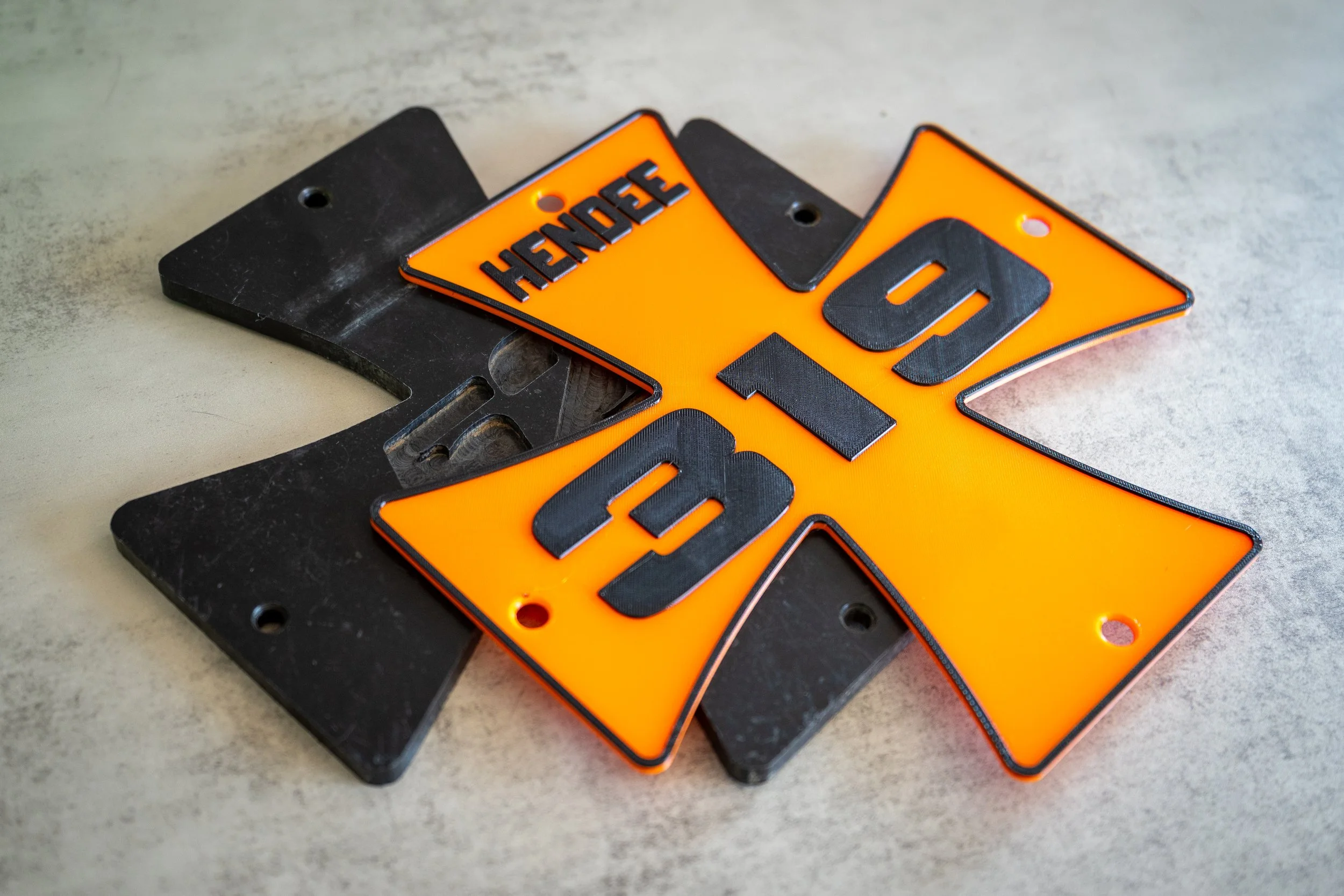 An original black plastic number plate, with a custom orange number plate made specifically for a customer.