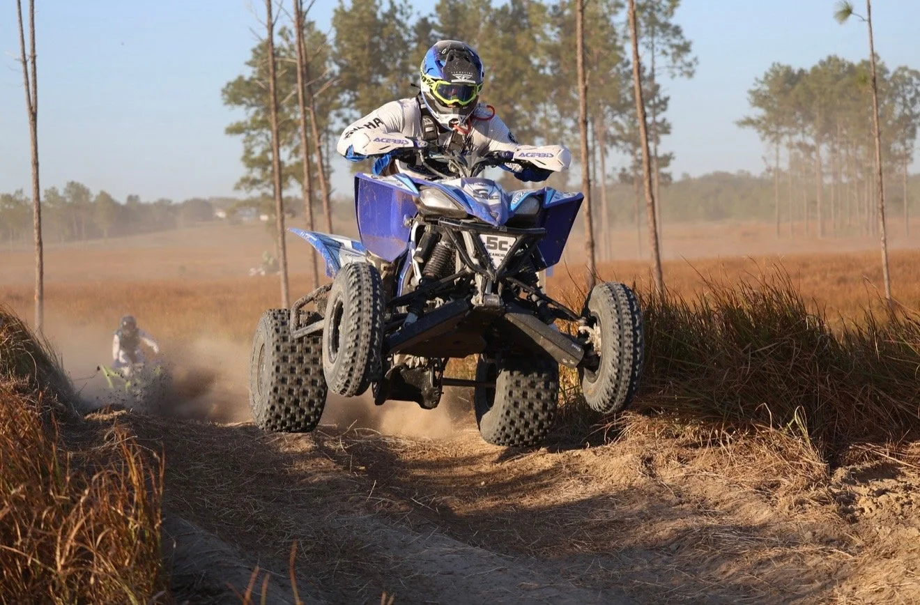 Helmeted rider jumps a blue ATV over a dirt trail in a pine forest.