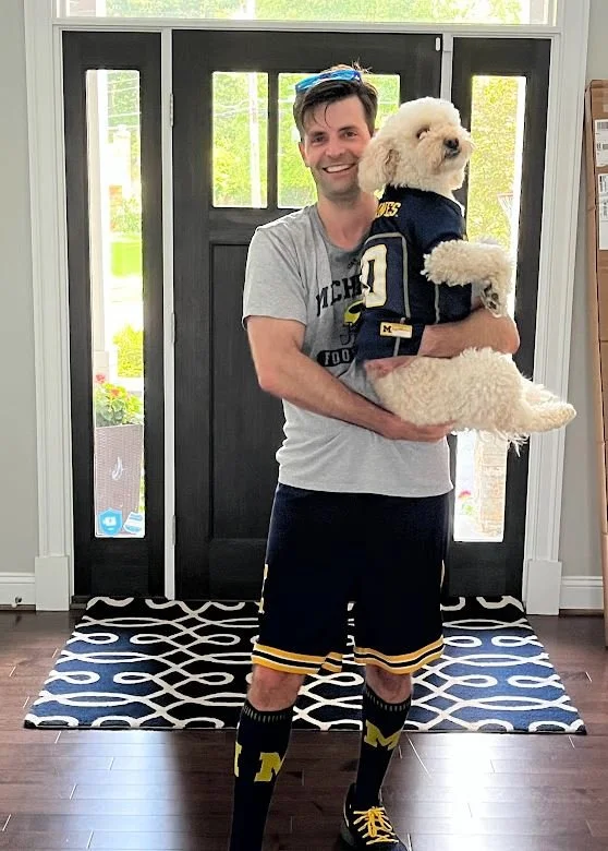 Dr. G holding Stella, a large, fluffy white dog dressed in a University of Michigan football jersey, standing inside a home with a front door and a black-and-white patterned rug.