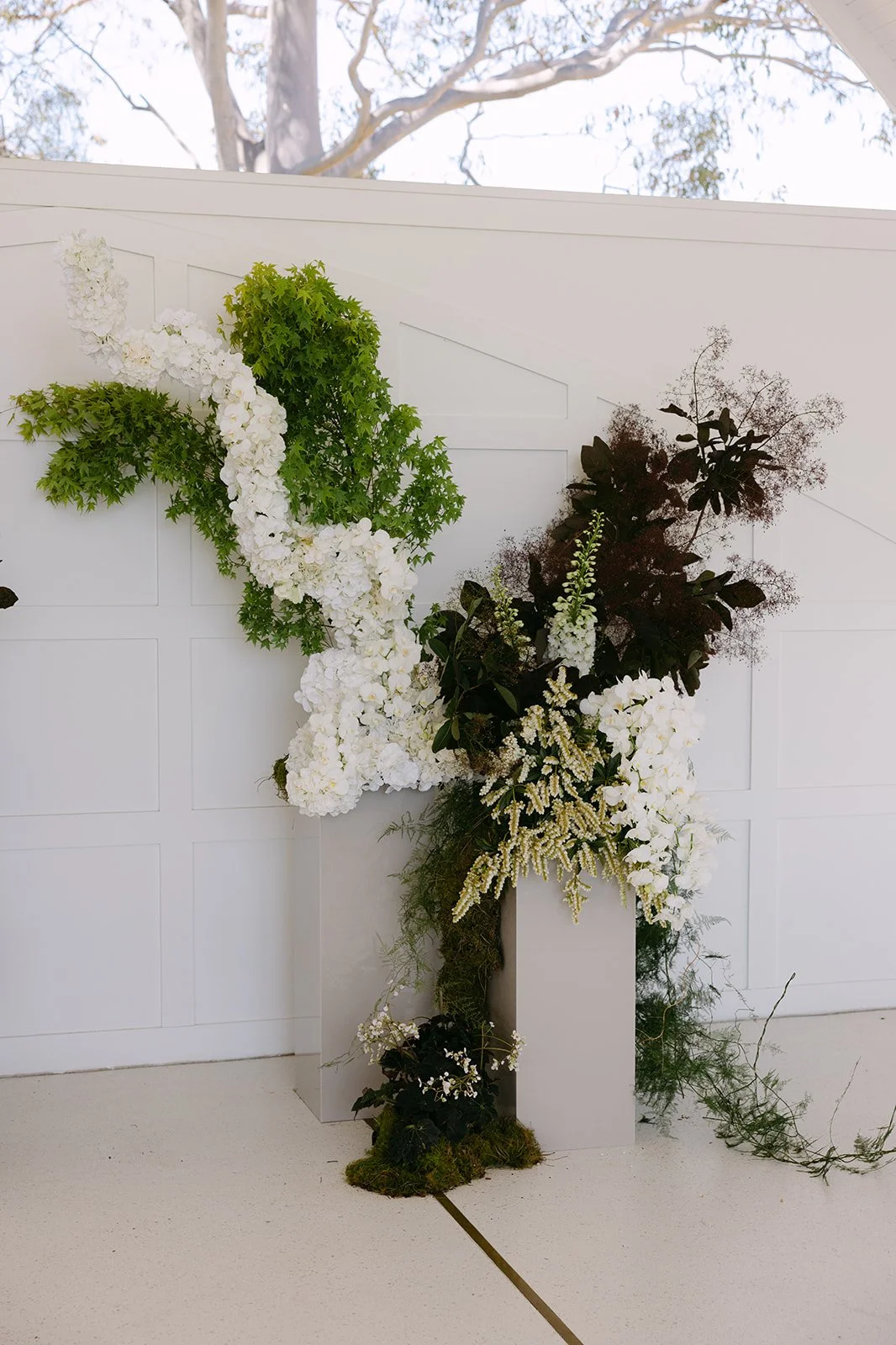 Floral arrangement with white and green flowers and dark leaves against a white wall.