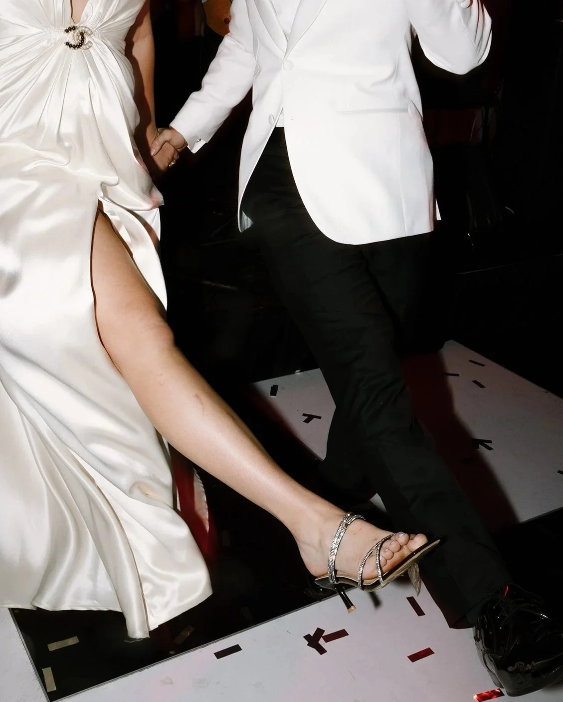 A couple holding hands, dressed in formal attire, with the woman's leg extended, revealing a high-heeled sandal, during a dance or special event.