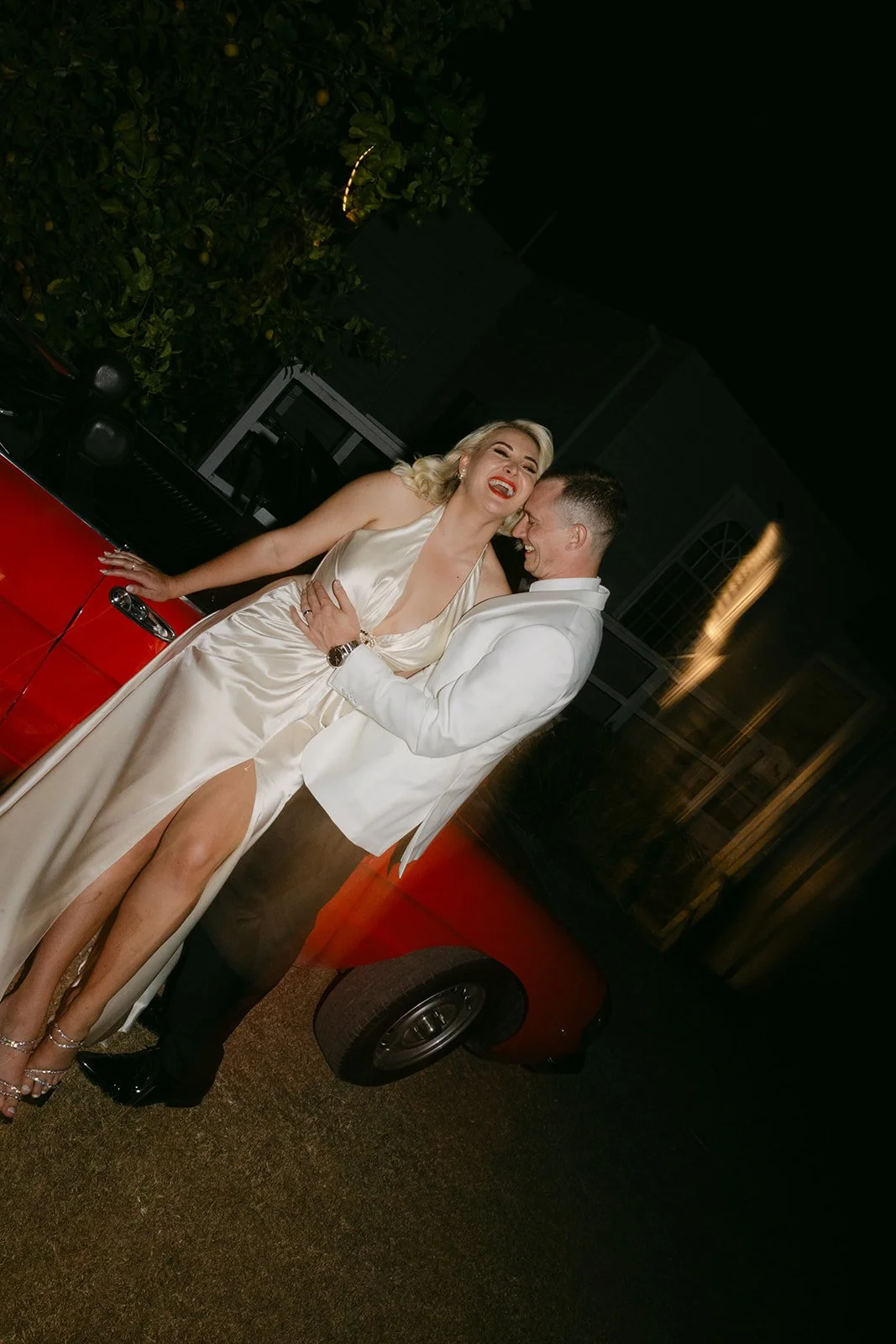 A couple in formal attire laughing and dancing next to a red vehicle at night.