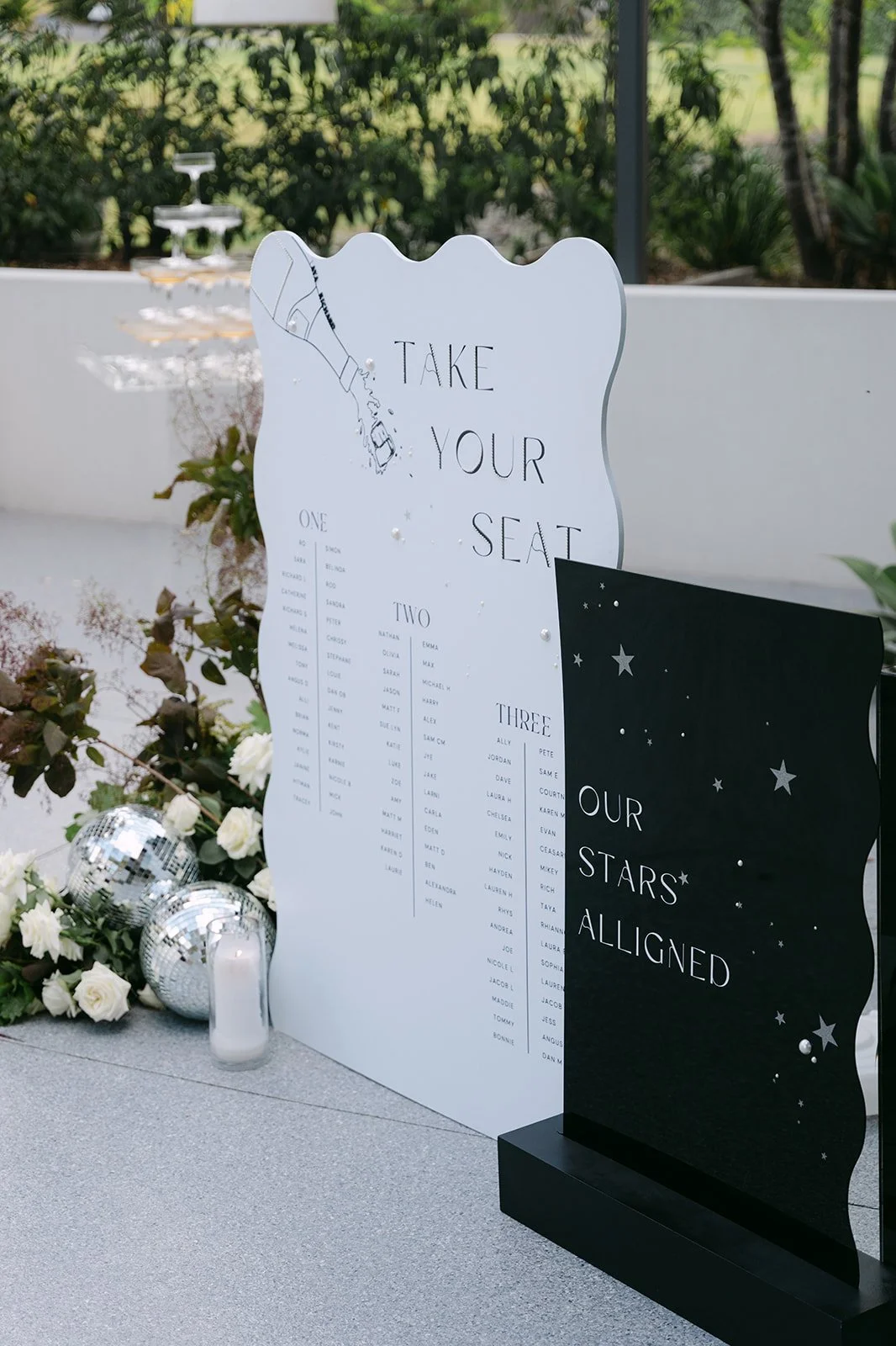 A seating chart at a wedding or event with a sign that says 'Take Your Seat'. The chart is decorated with illustrations of a champagne bottle pouring into a glass and a starry night sky with the phrase 'Our Stars Aligned'. There are white roses, disc