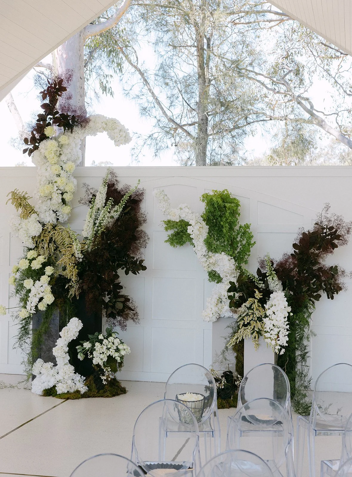 Floral wedding arch with white, green, and dark purple flowers, set against a white wall with an outdoor view of trees and blue sky.
