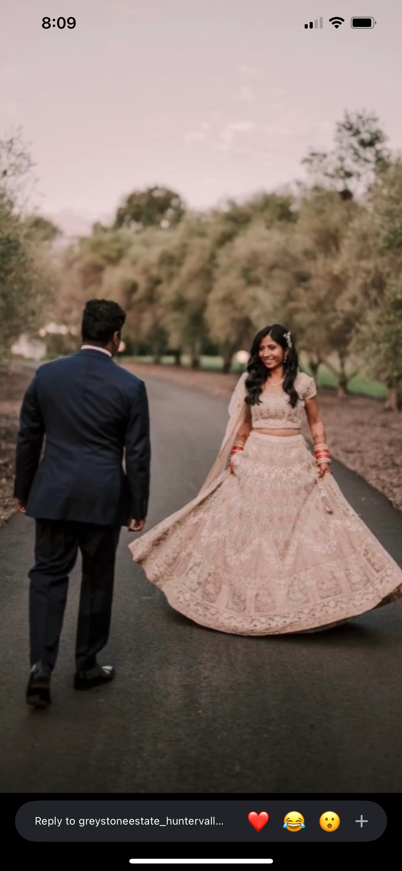 A bride and groom walking on a paved path in a park with trees, the bride in a traditional Indian dress and the groom in a dark suit.