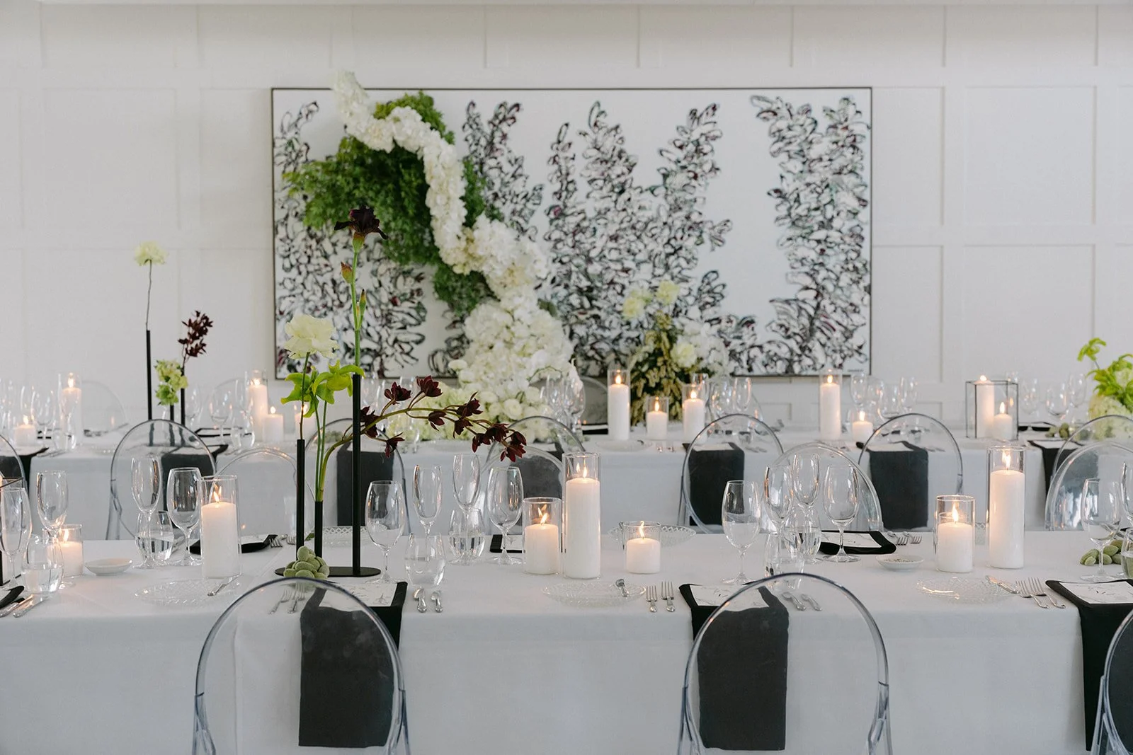 Elegant banquet table set with candles, flowers, and glassware, decorated with a black and white theme, in front of an abstract black and white floral artwork.