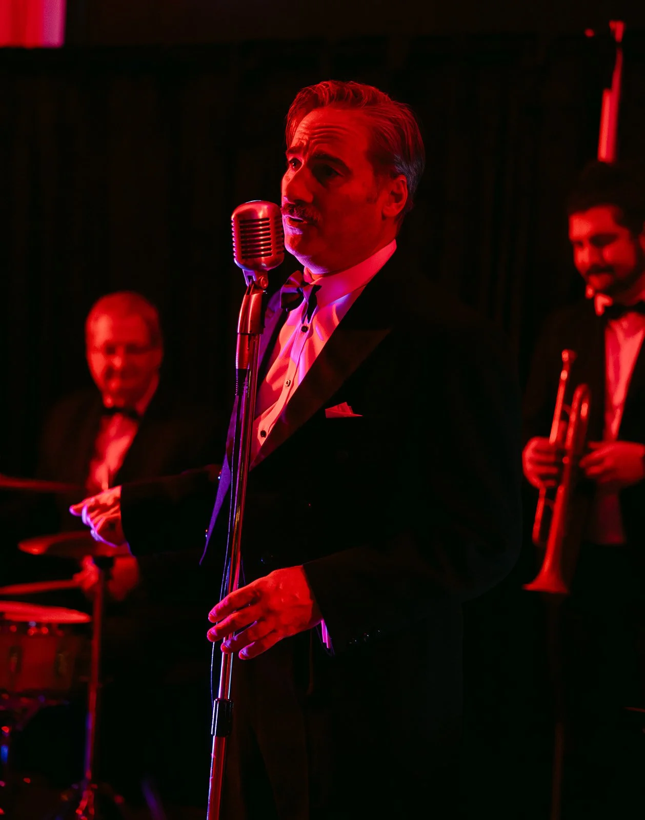 A male singer in a tuxedo performing on stage with a vintage microphone, accompanied by a jazz band with a drummer and a trumpet player, all dressed formally in black suits and bow ties, under dim red lighting.