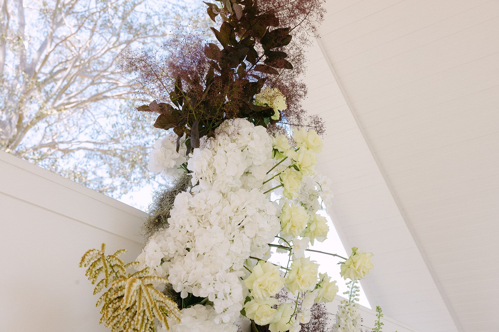 A floral arrangement of white and cream flowers with dark green and purple leaves, hanging from the ceiling against a white interior wall.