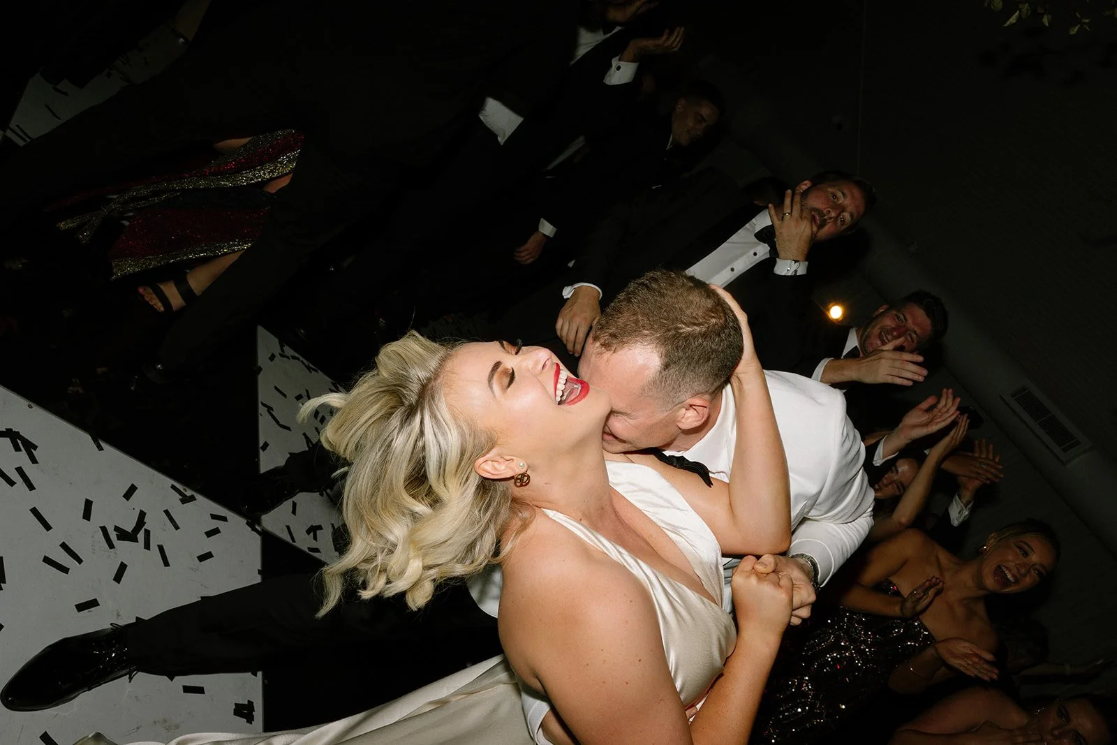 A woman and a man share a kiss at a celebration, surrounded by friends clapping and smiling, with confetti on the floor.