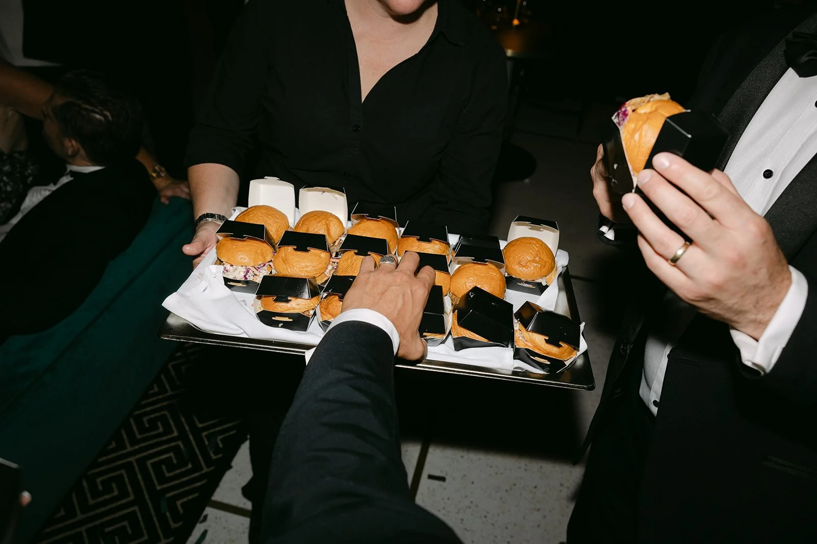 A person in a tuxedo holding a slider bun, another person in black attire holding a tray with small sliders, and a woman in black serving others at a social event.
