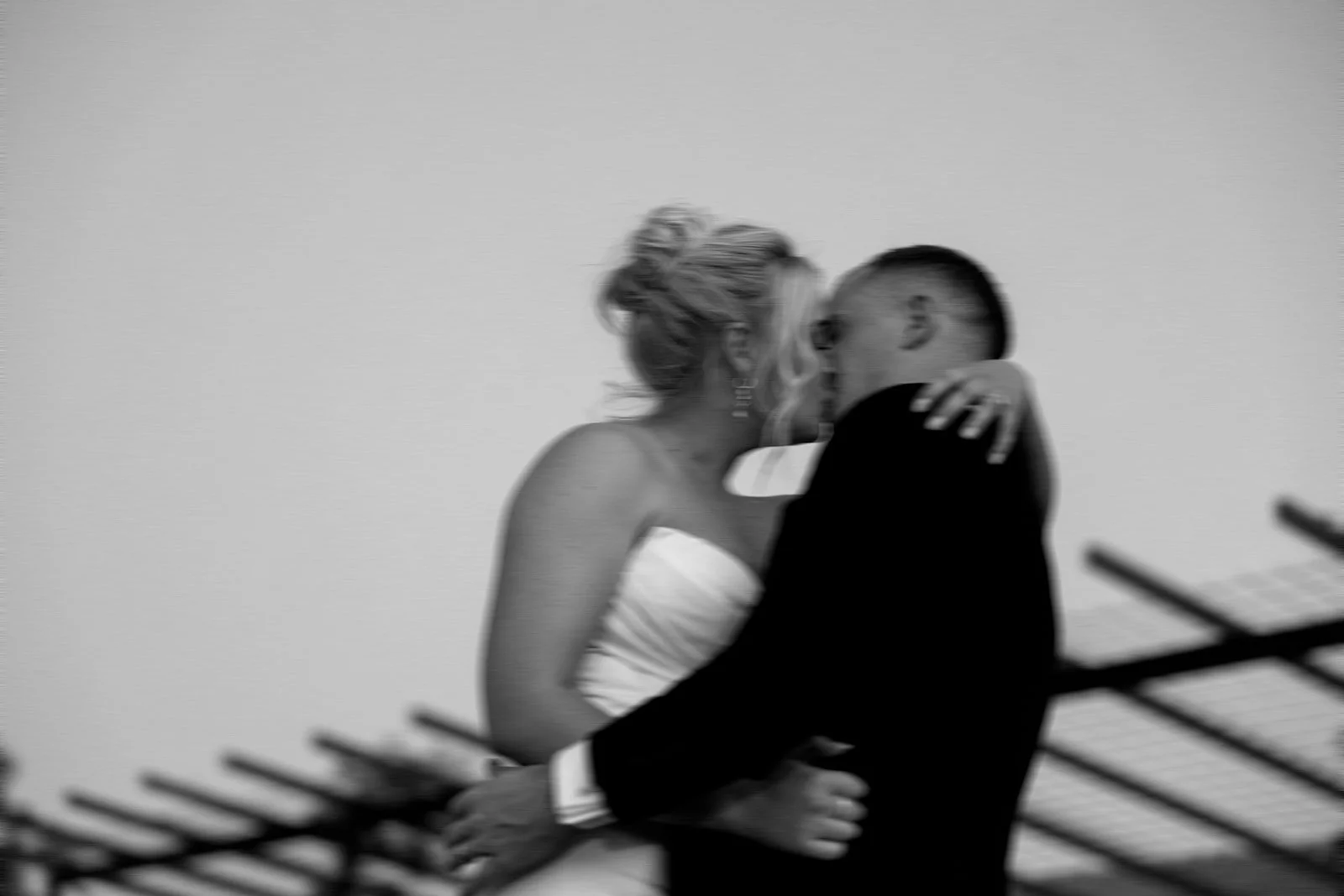Black and white photo of a couple kissing, with the woman wearing a strapless dress and the man in a suit, behind a fence.