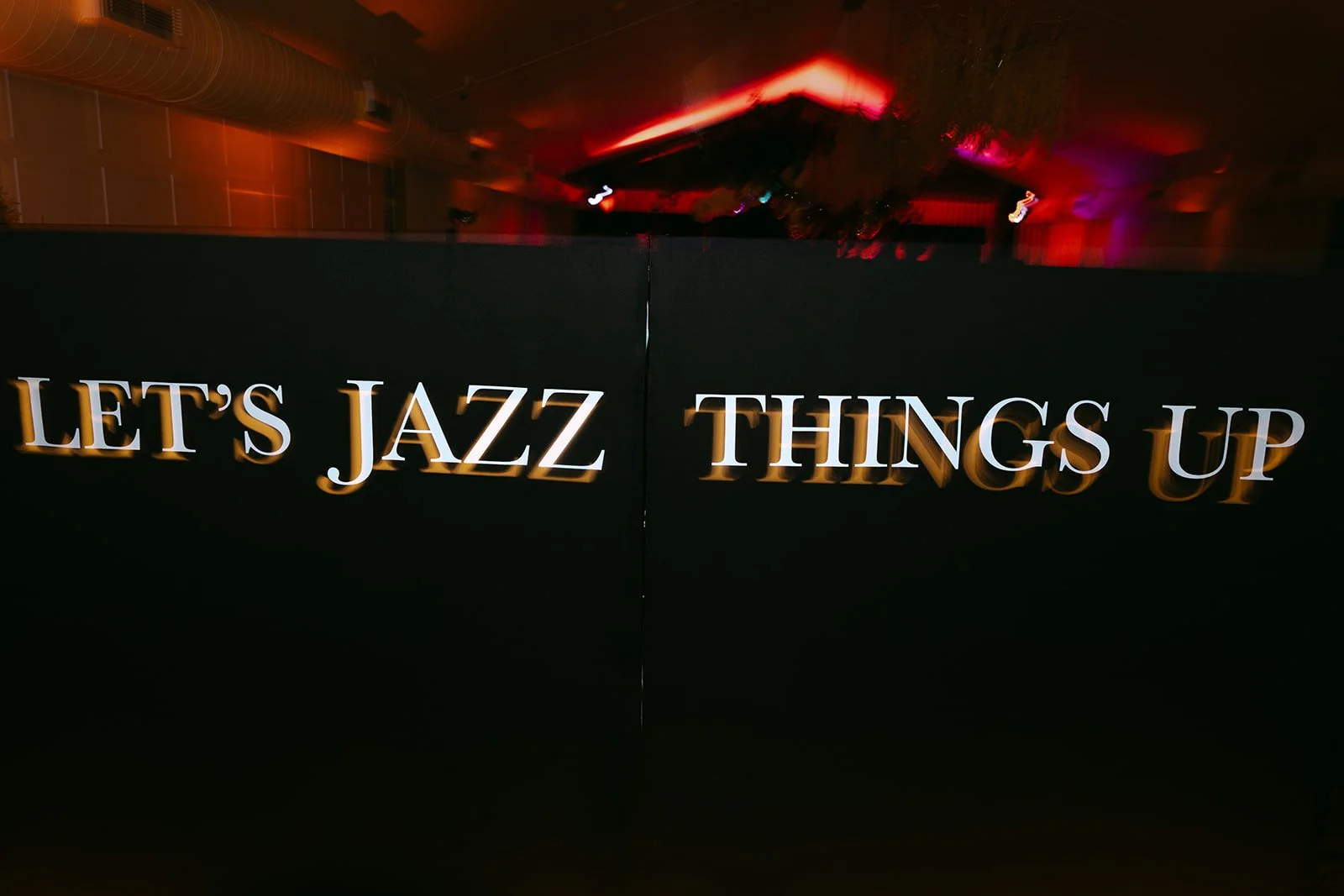 A black divider or wall with illuminated white text that says "LET'S JAZZ THINGS UP" in a ballroom or event space with dim lighting and red and purple accent lights.