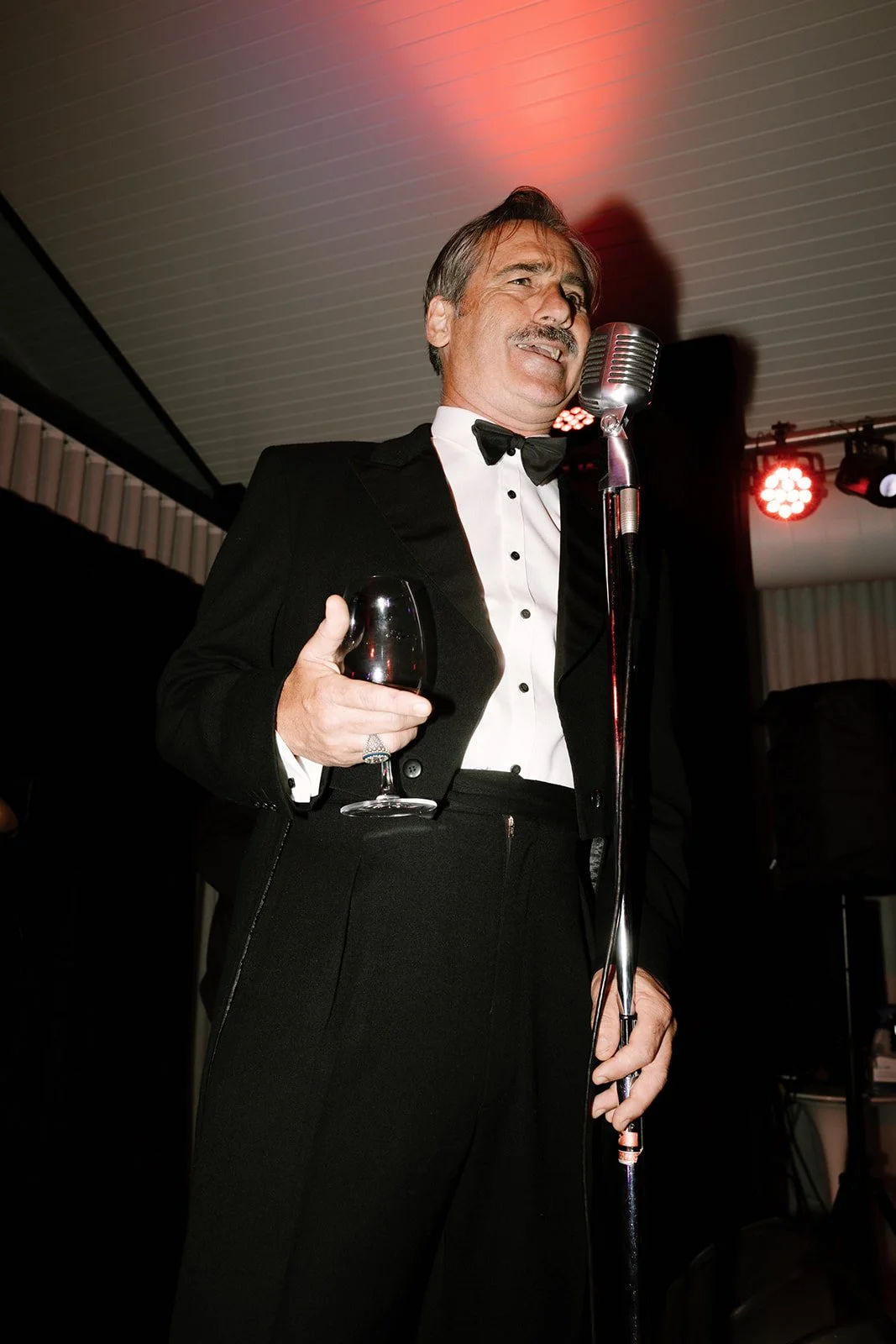 Man in a tuxedo holding a glass of red wine, standing in front of a vintage microphone on stage.