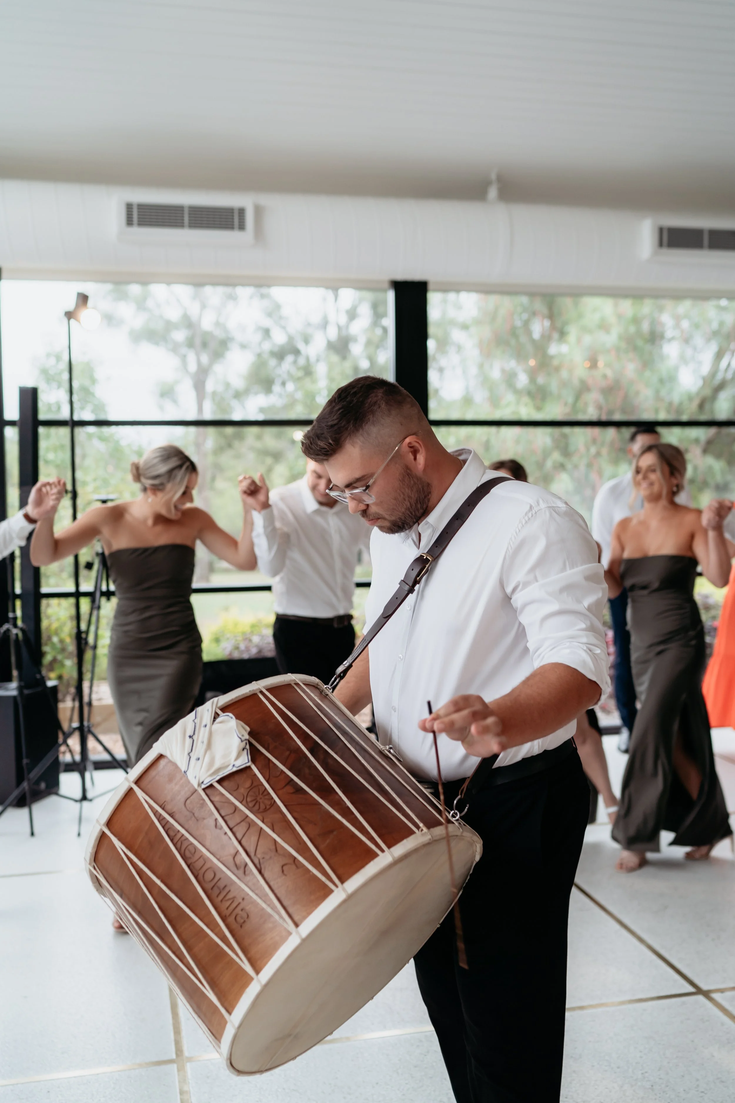 A man playing a traditional drum at a lively celebration, with women dancing in the background.
