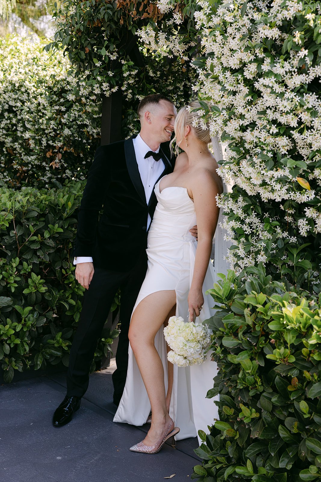 A newlywed couple smiling and leaning close to each other outdoors surrounded by white flowers and green foliage. The bride is holding a bouquet and wearing a strapless white dress with a slit, while the groom is in a black tuxedo with a bow tie.