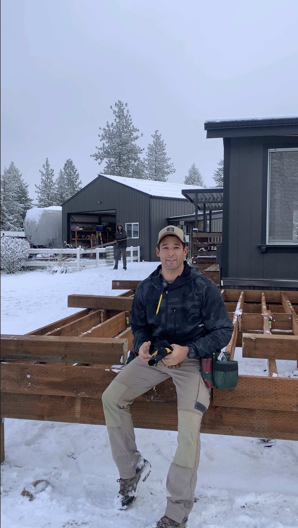 A man sitting on a wooden frame outdoors in snowy weather, with a dark gray house to the right and a shed with oriented strand board in the background, along with another person standing near the shed.