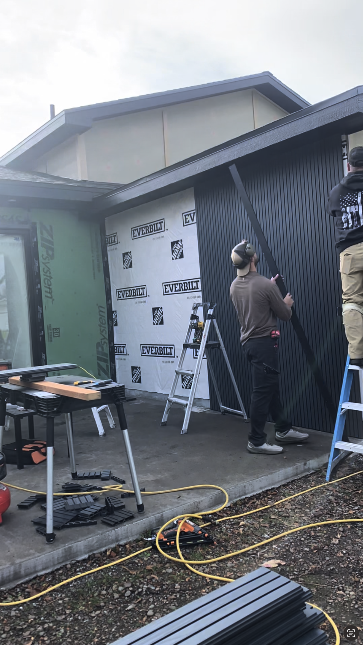 People working on installing black vertical siding on a house exterior. There are ladders, tools, and construction materials around, with wall insulation labeled 'Everbilt' and 'Zip System' visible.