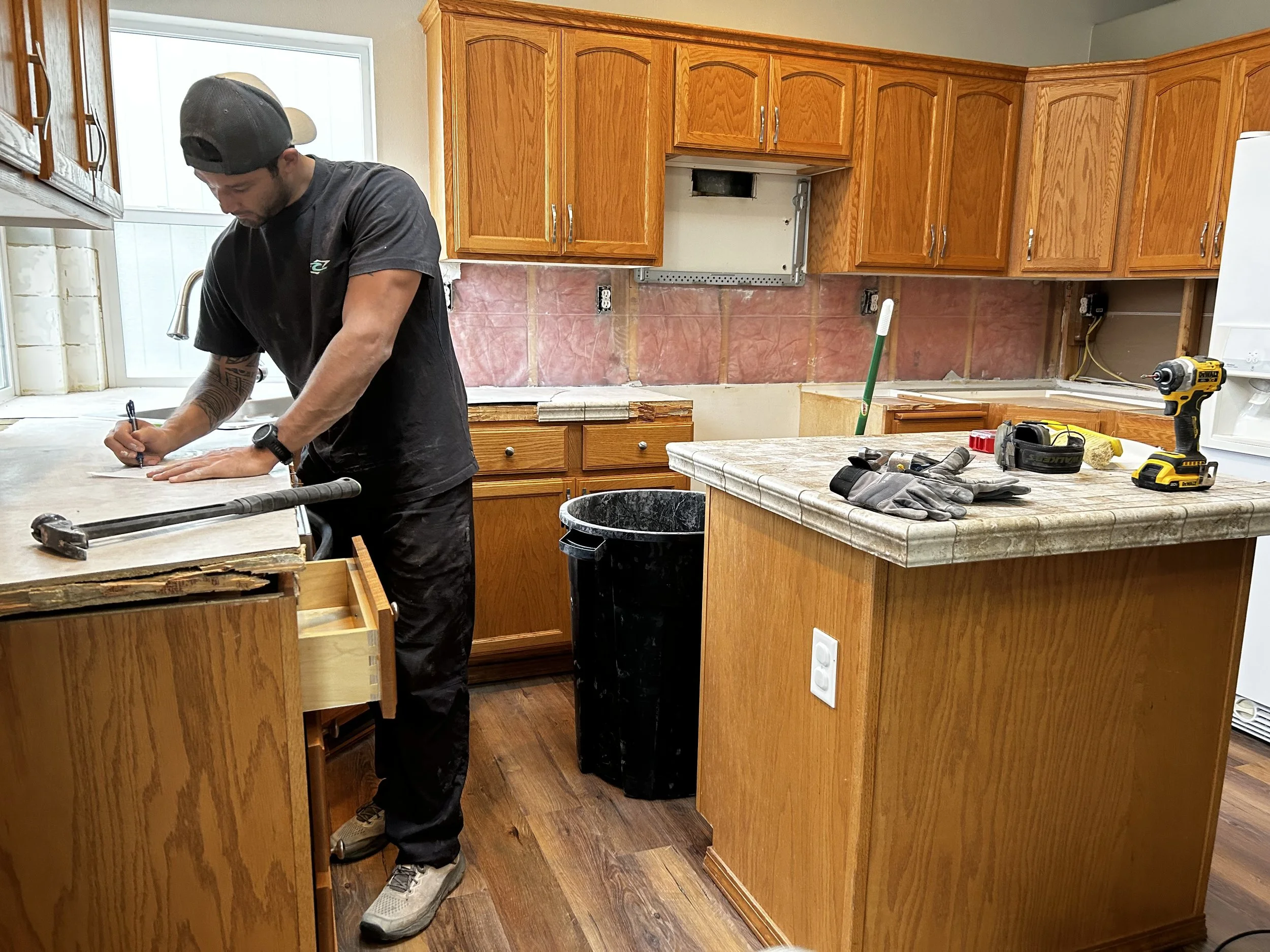 Man working on kitchen countertop renovation with tools and gloves on island counter, unfinished wall with drywall and cabinet installation.
