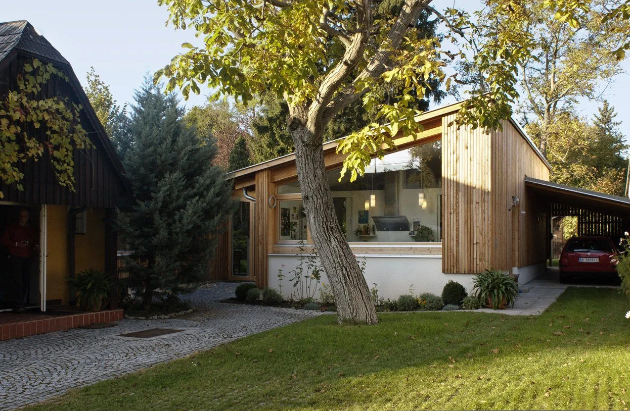 A modern house with large glass windows and wooden siding, surrounded by trees and a well-maintained lawn, with a red car parked on a driveway.
