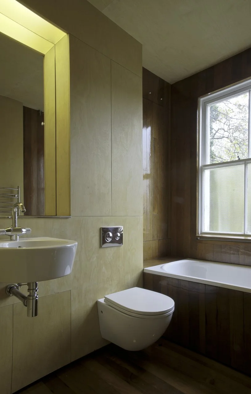 A bathroom with beige tiled walls, a small wall-mounted sink, a wall-mounted toilet, a bathtub by a large window, and wooden flooring.