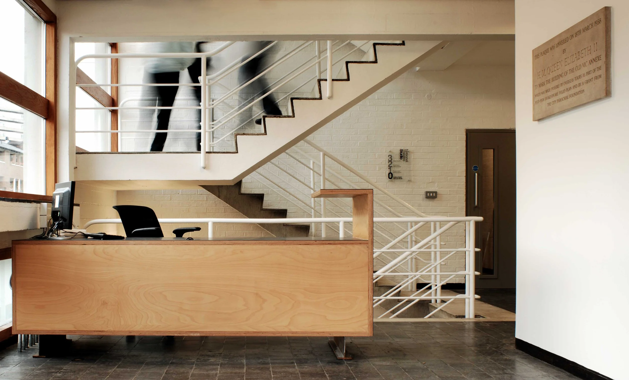 Office reception area with a wooden desk, computer, black chair, and a staircase with white railings leading upstairs, behind which a person is walking.