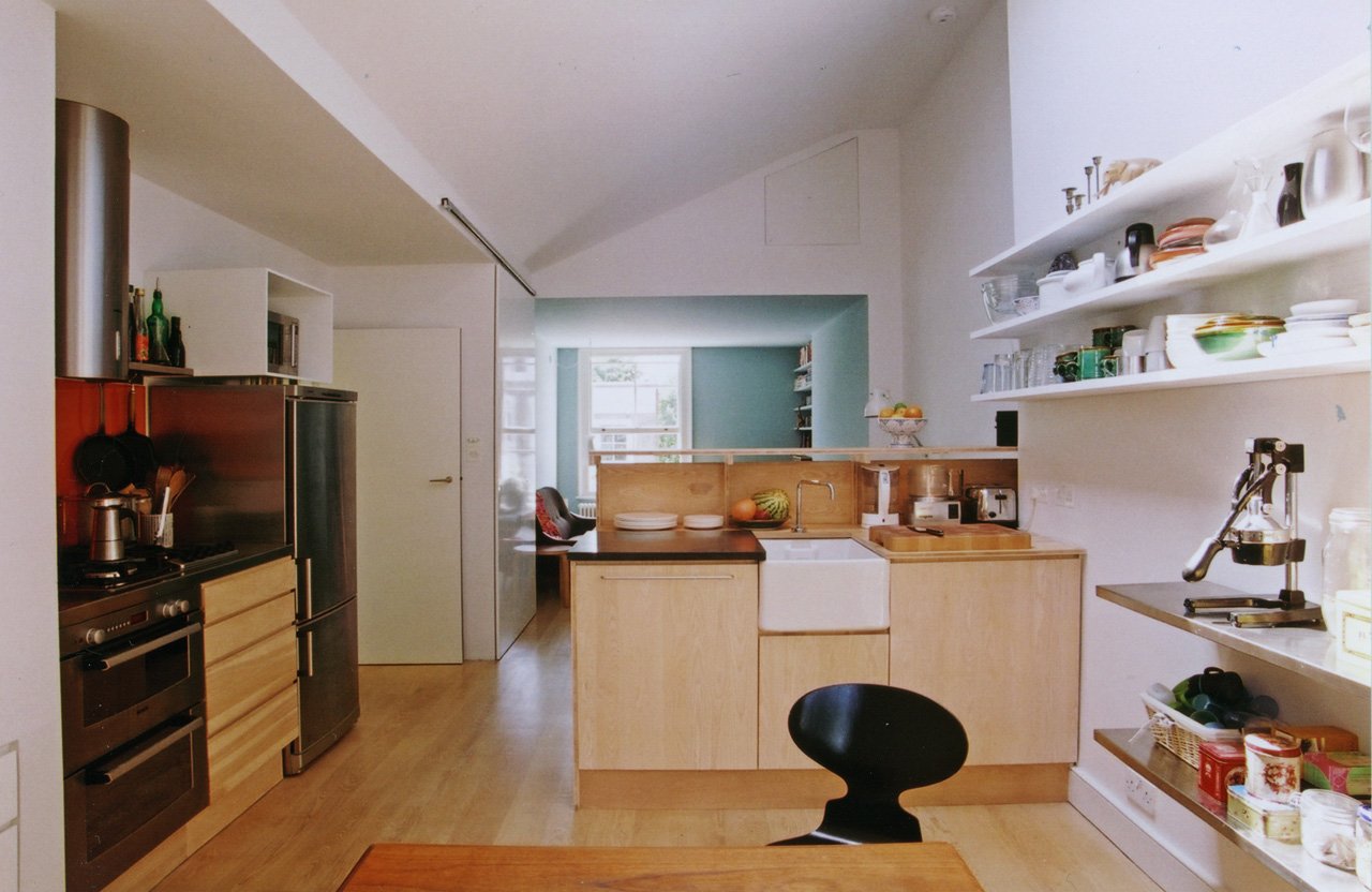 Interior of a modern kitchen with wooden cabinets, open shelves, and a white farmhouse sink. There are various kitchen appliances, dishes, and ingredients visible, with a seating area in the background.