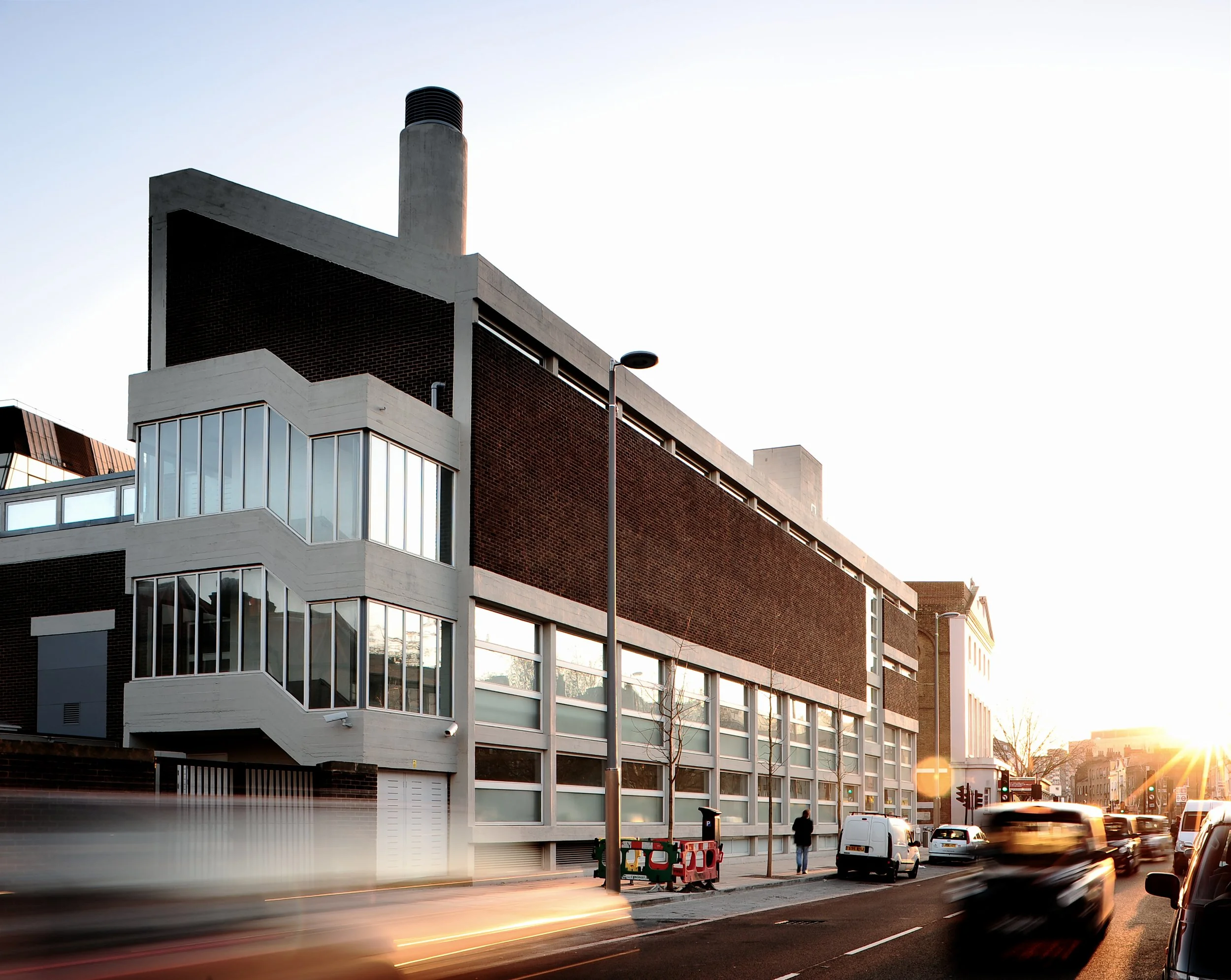 Modern multi-story building with large windows and a brick facade at sunset, with cars and pedestrians on the street.