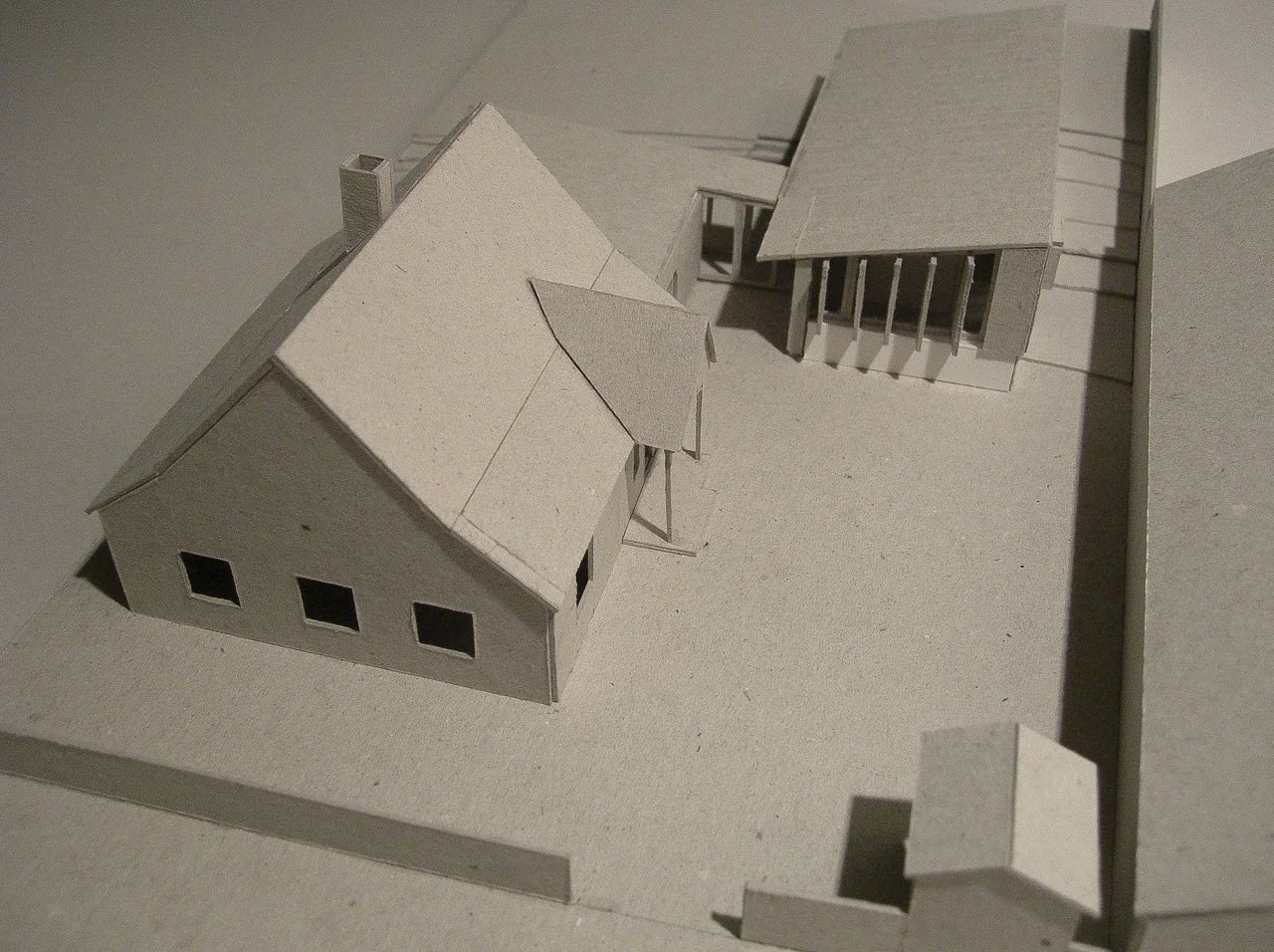 Architectural scale model of a residential property with multiple buildings, including a main house with three windows, a detached garage or shed, and an additional structure, all set on a flat base.