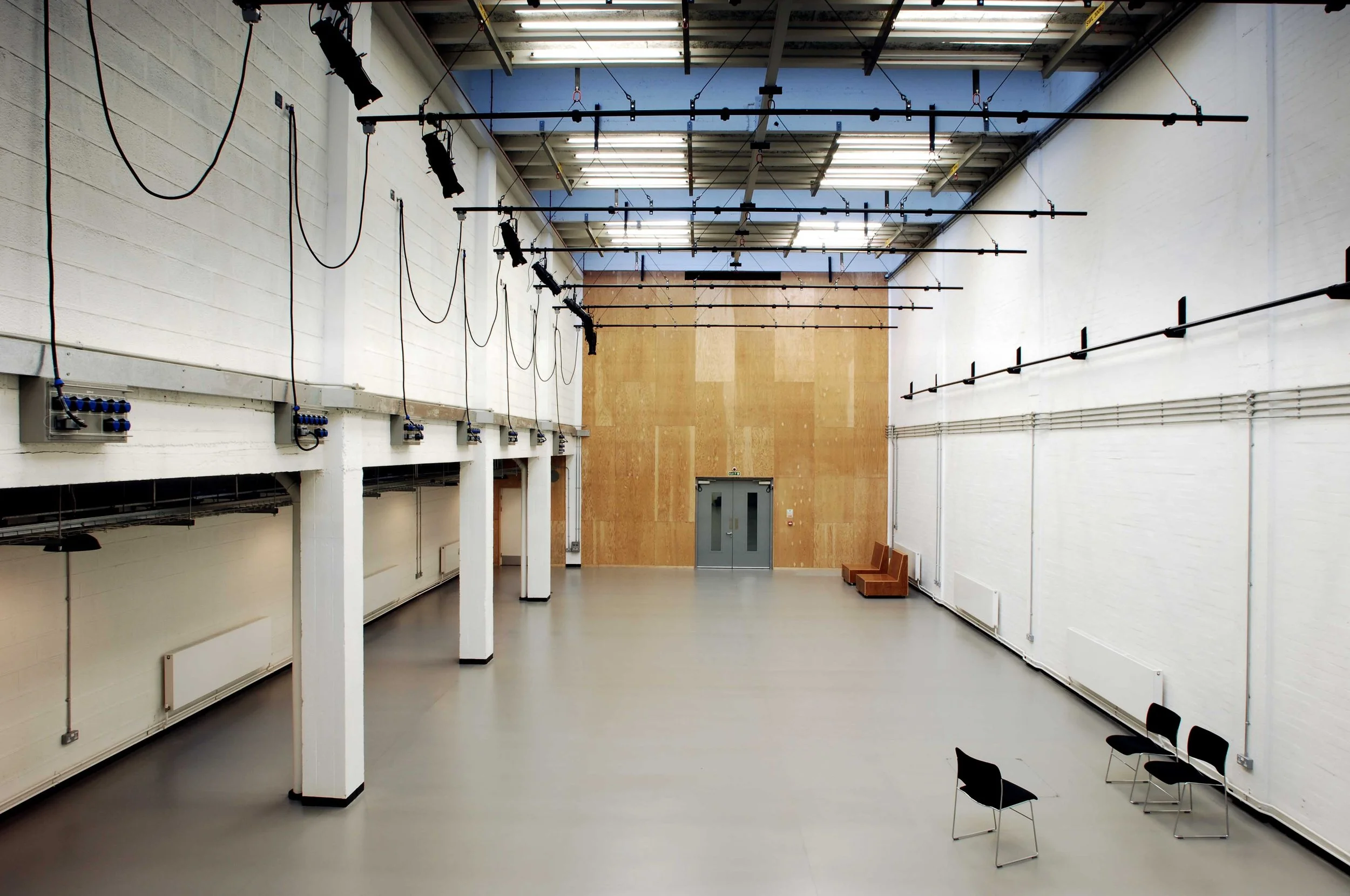 Empty rehearsal or performance studio with black chairs, white walls, a wooden partition, and stage lighting and equipment hanging from the ceiling.